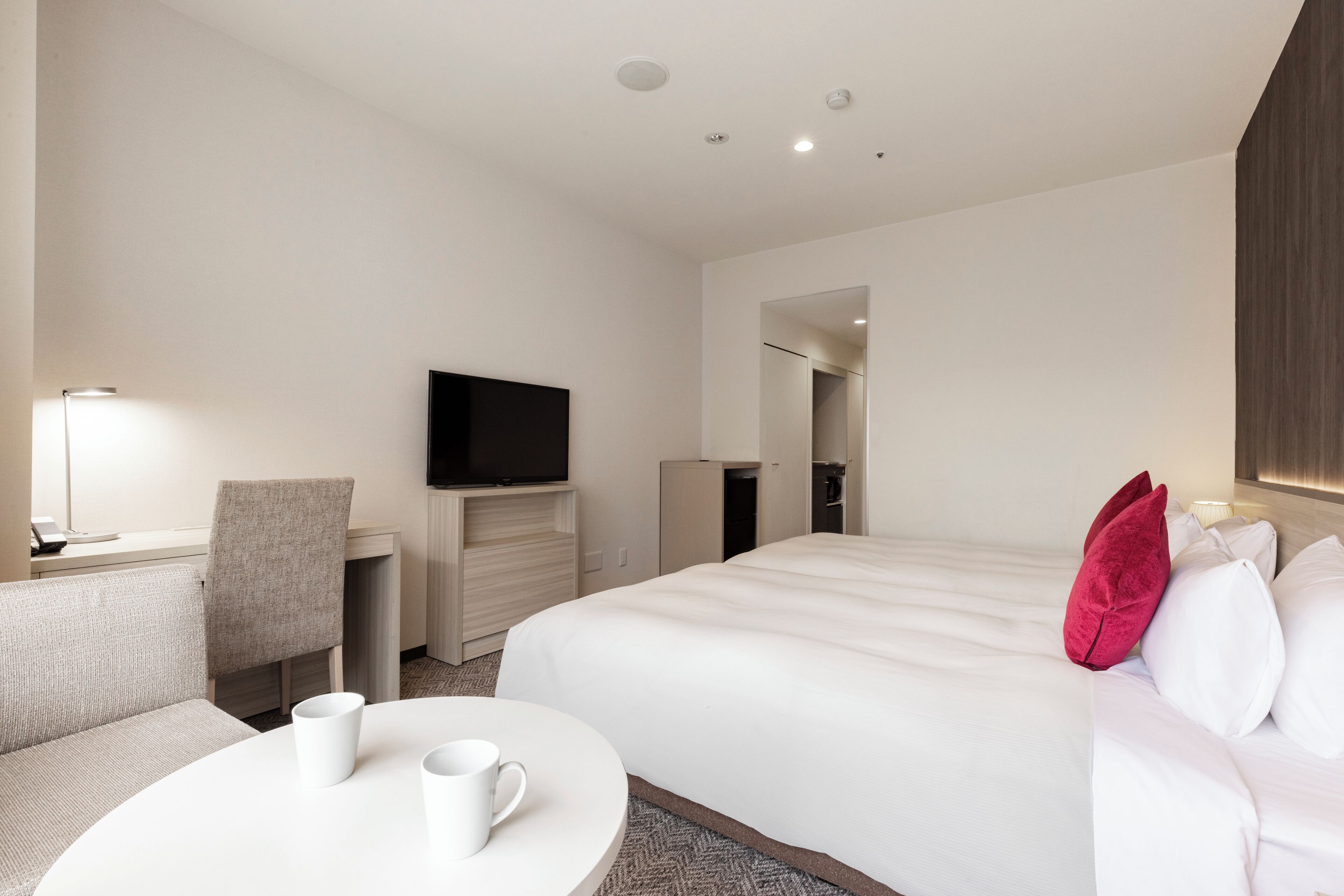 A hotel room with a bed, a desk with a flat-screen TV, a chair, and a round table with two white mugs.