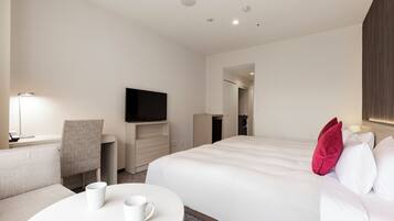 A hotel room with a bed, a desk with a flat-screen TV, a chair, and a round table with two white mugs.