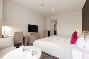 A hotel room with a bed, a desk with a flat-screen TV, a chair, and a round table with two white mugs.