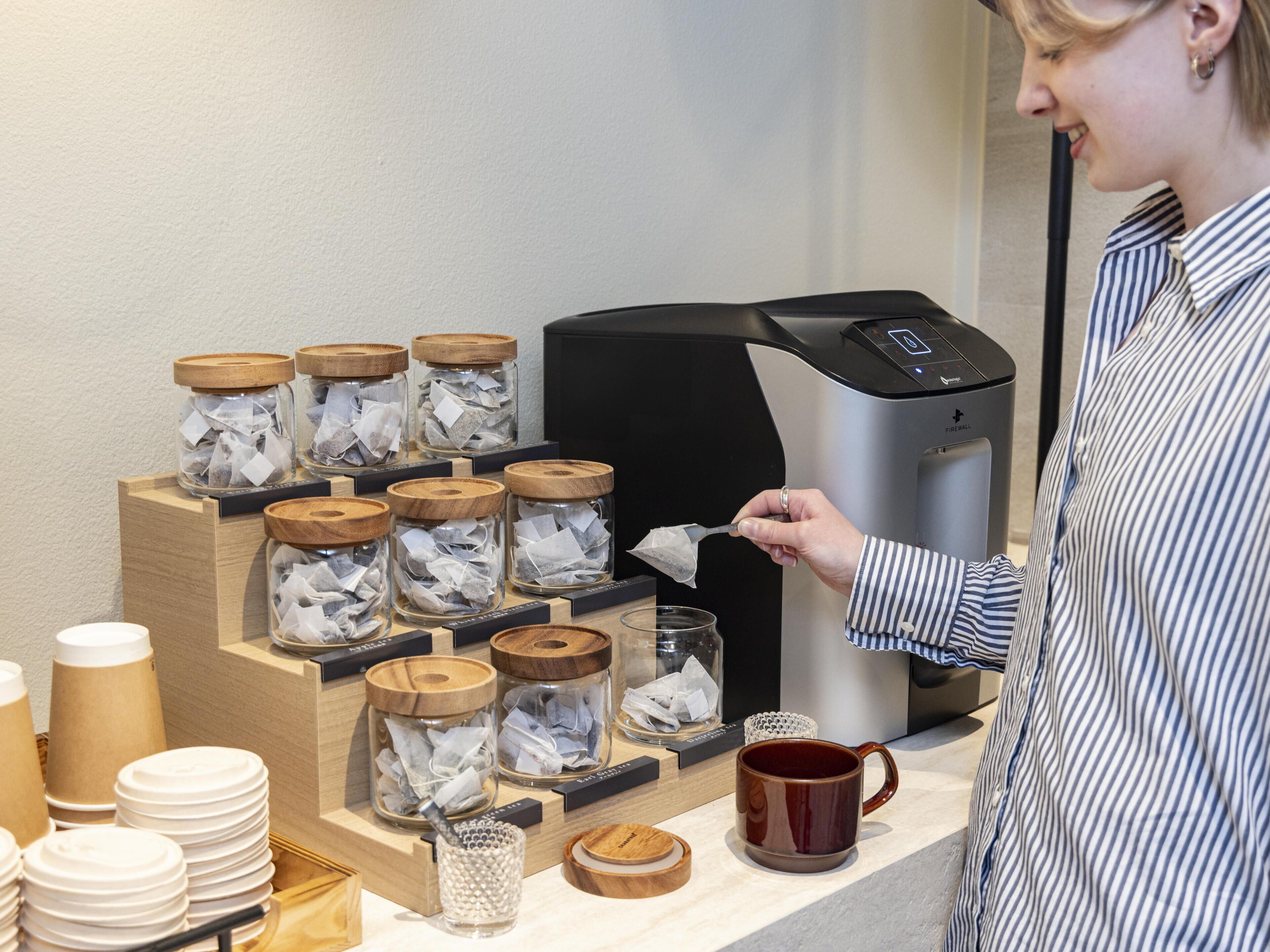 A coffee station with a coffee machine, tea dispensers, and stacked paper cups.
