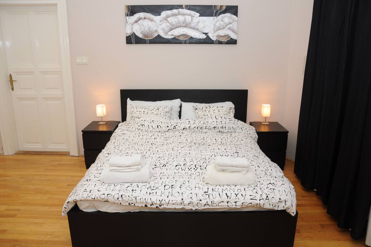 Una cama cuidadosamente tendida con una colcha de diseño alfabético blanco y negro, dos mesitas de noche con lámparas, y una obra de arte en la pared sobre la cama.