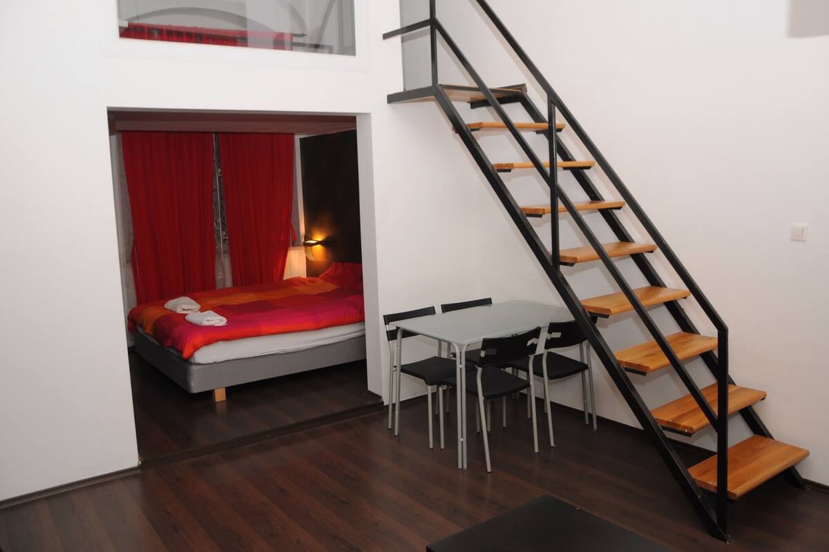 Una habitación pequeña con una cama, una mesita y una escalera.