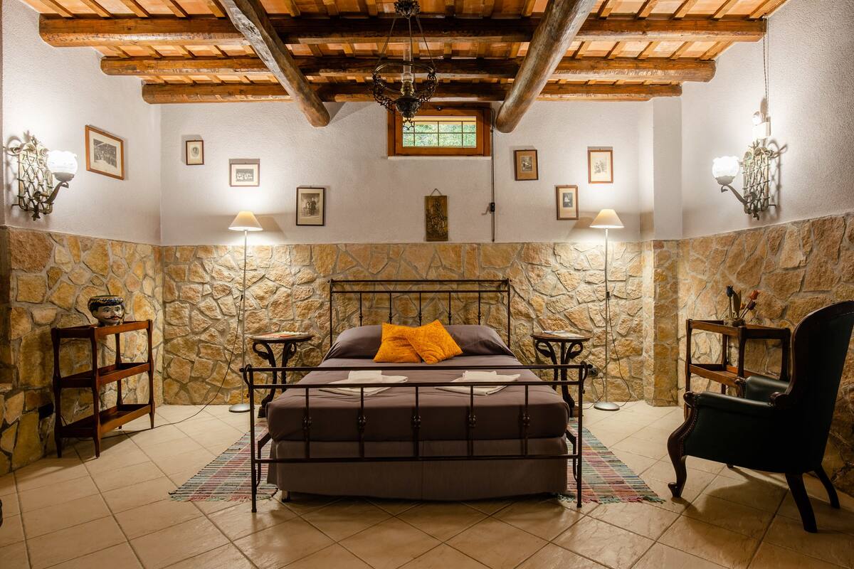 A bedroom with a stone wall, a bed with a metal frame, two bedside tables, a chair, and a wooden ceiling.
