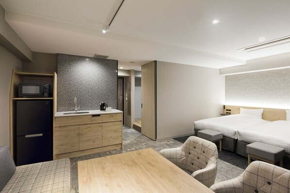 Une chambre d’hôtel moderne équipée d’un lit, d’un petit bureau, d’un four à micro-ondes, d’une armoire en bois et d’un coin salon avec deux fauteuils.