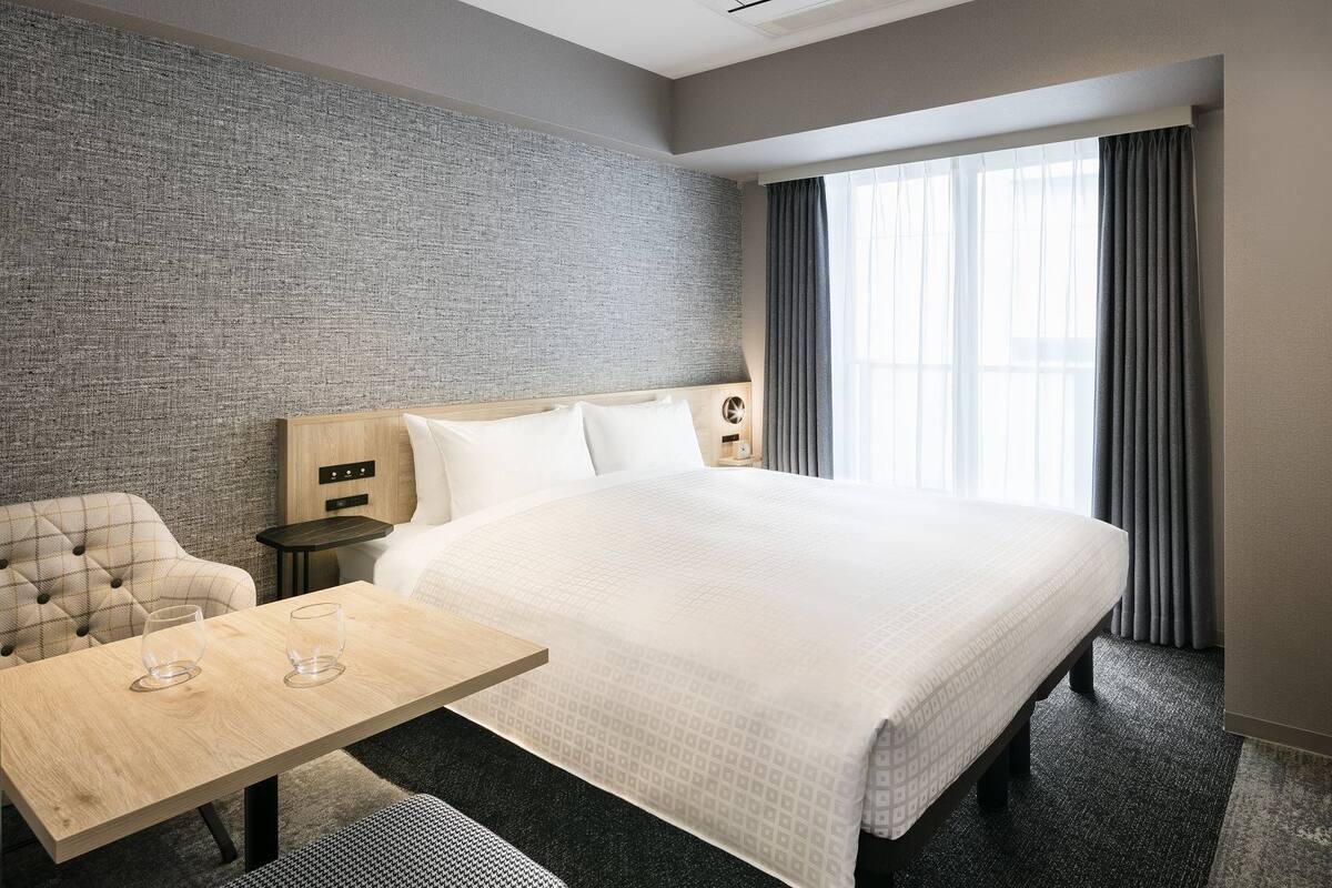 A hotel room with a large bed, a wooden nightstand, a desk with two glasses, a chair, and a television.