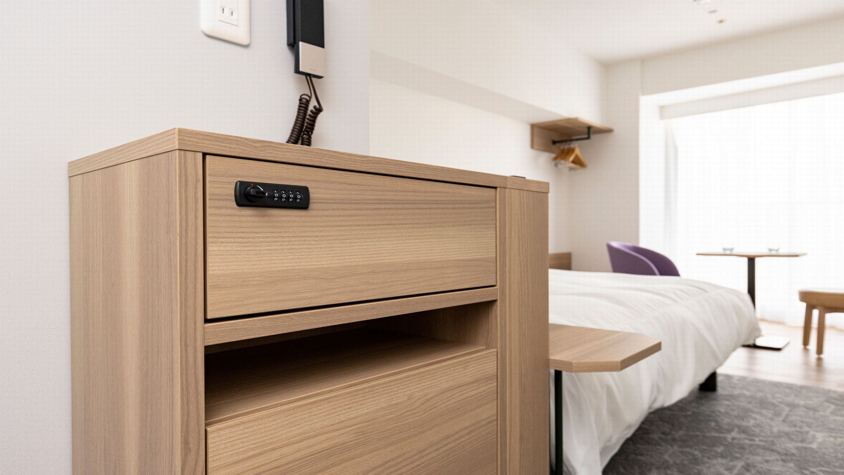 A wooden bedside table with a control panel, a bed with white bedding, a small table with two cups, and a purple chair in a hotel room.