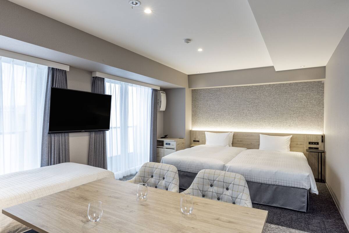A modern hotel room with two beds, a flat-screen TV, a dining table, and a large window with curtains.