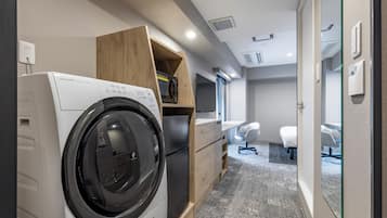 Ein kleines Hotelzimmer mit Waschmaschine, einer kleinen Holzablage, einem Schreibtisch, einem Stuhl und einem Bett.