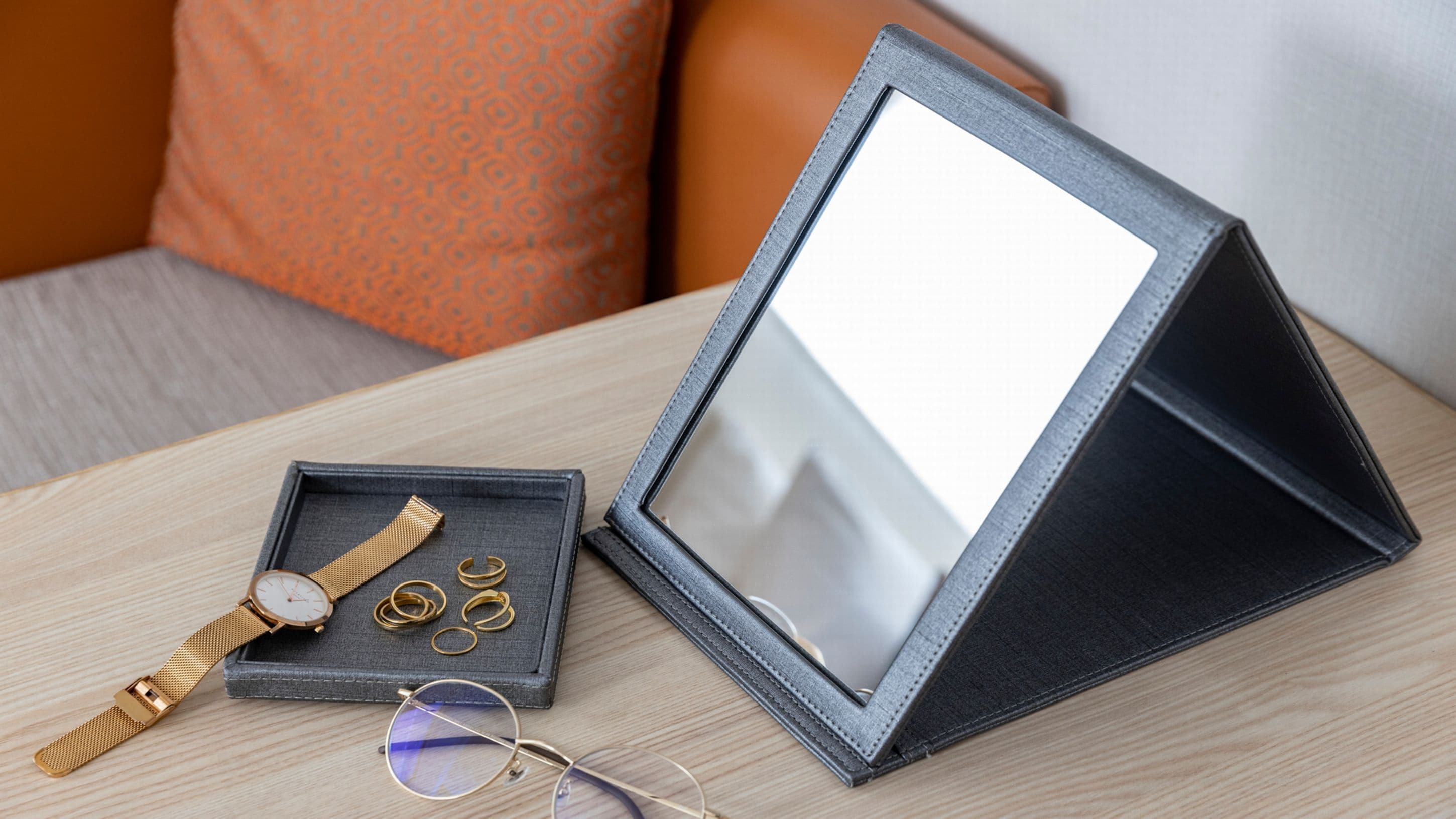 A black leather case with a mirror, a wristwatch, and gold rings on a wooden table.