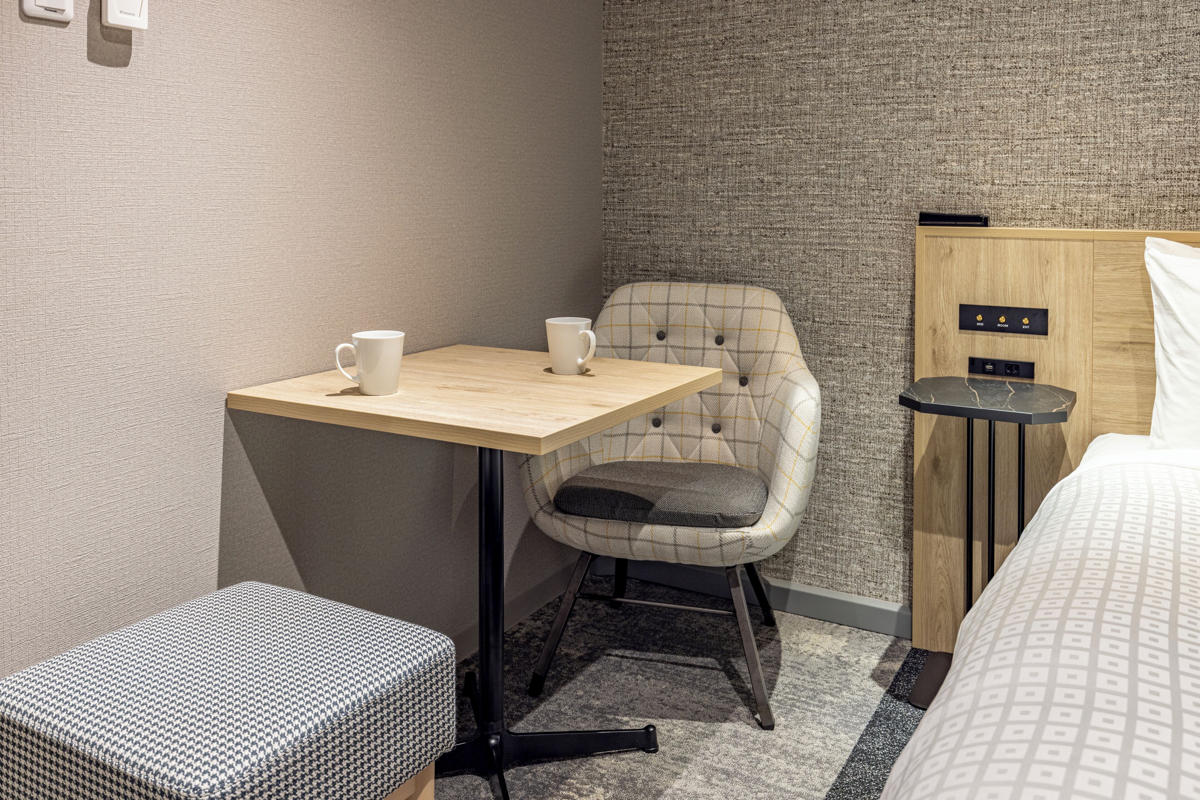 A small, neatly arranged hotel room with a wooden table, a plaid chair, a patterned ottoman, a bed with a patterned bedspread, and a textured wall.