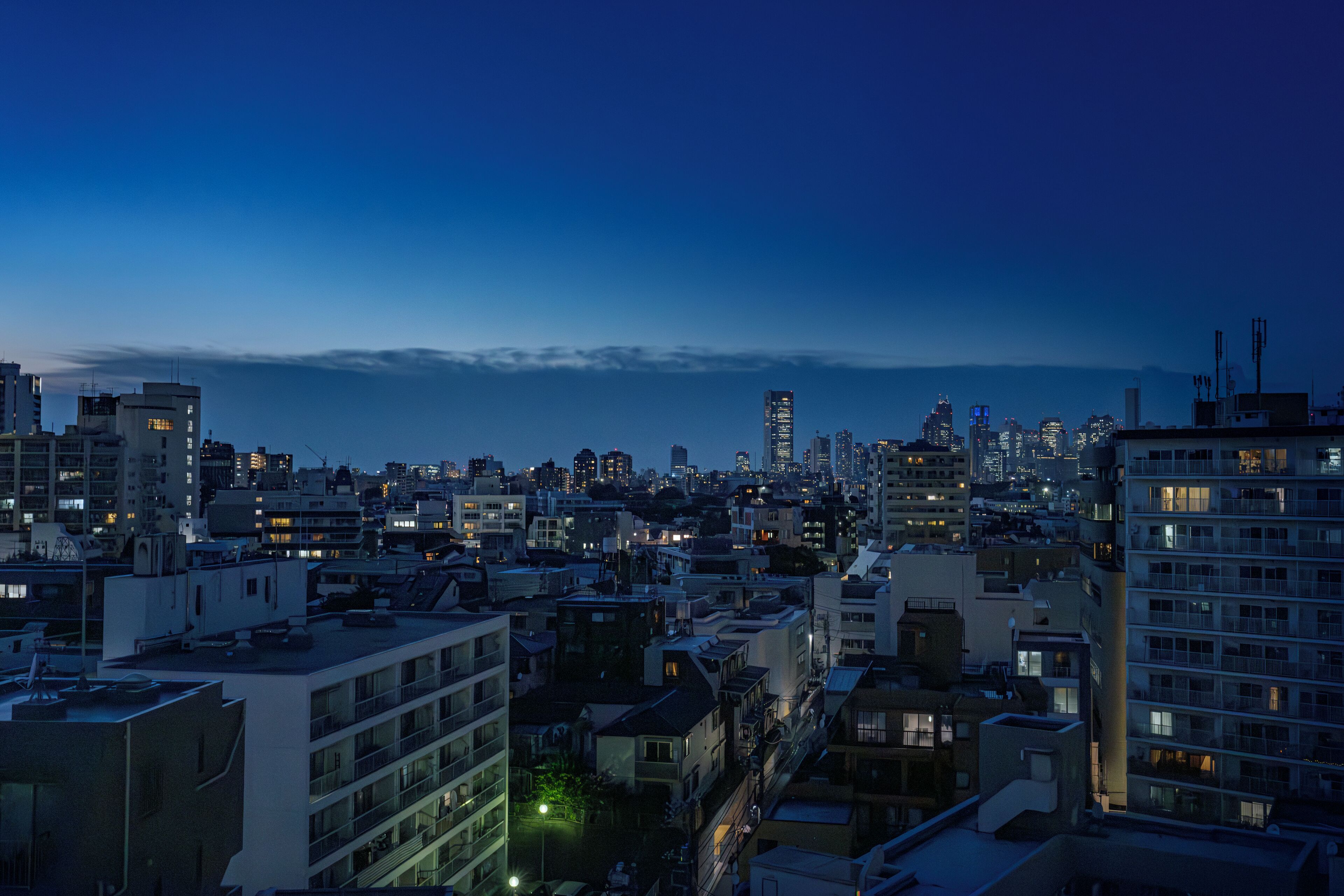 A cityscape at dusk with numerous buildings, some with visible balconies and windows, and a few with illuminated signs.