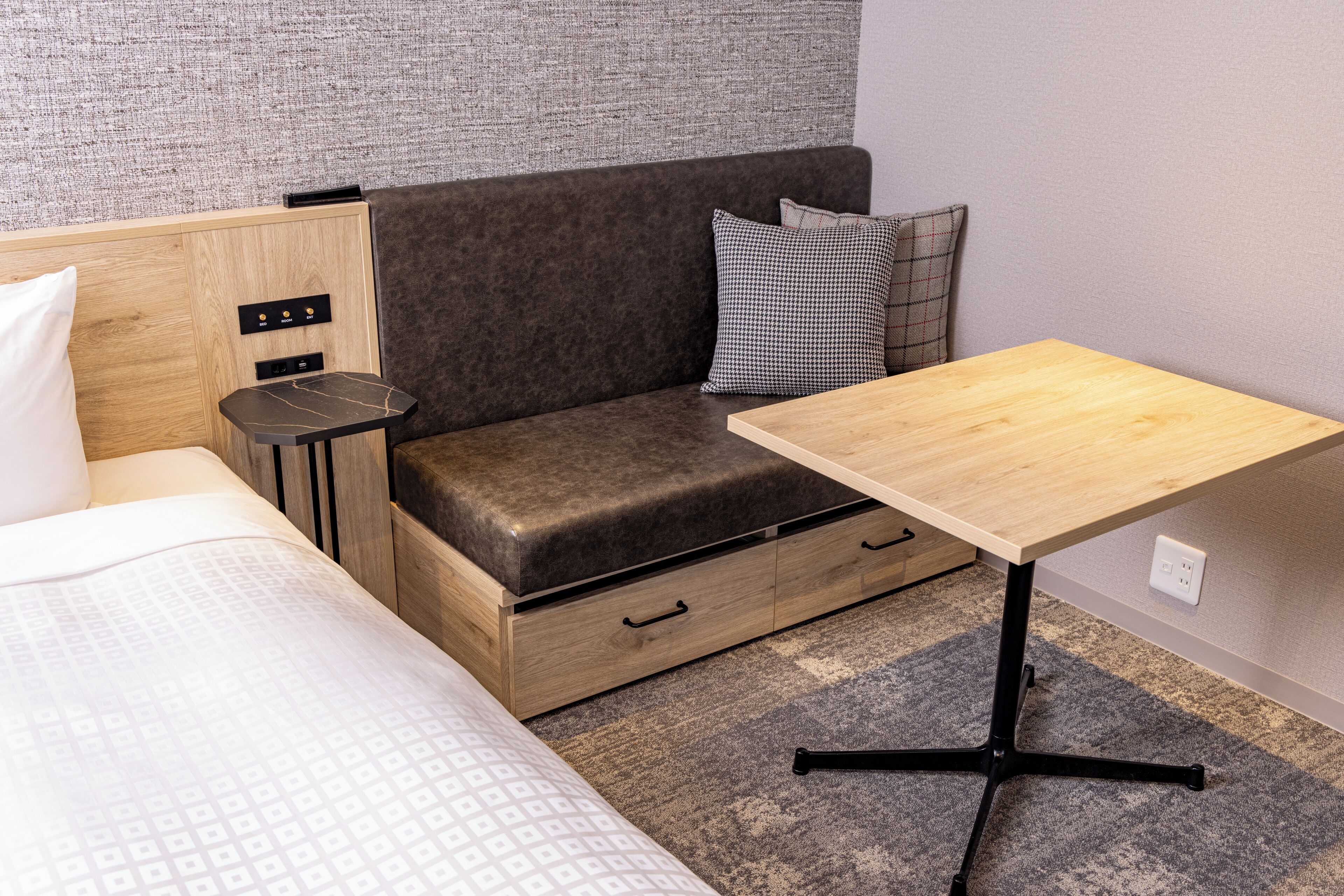A compact room with a bed, a small sofa with storage drawers, a wooden table, and a wall-mounted power outlet.