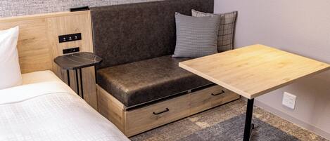 A compact room with a bed, a small sofa with storage drawers, a wooden table, and a wall-mounted power outlet.