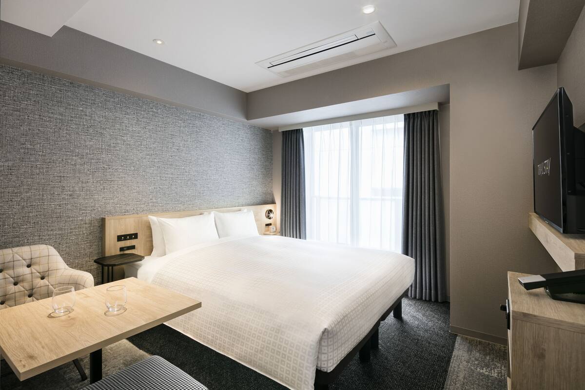 A modern hotel room with a large bed, a wooden headboard, a flat-screen TV, a small table with glasses, and a grey textured wall.