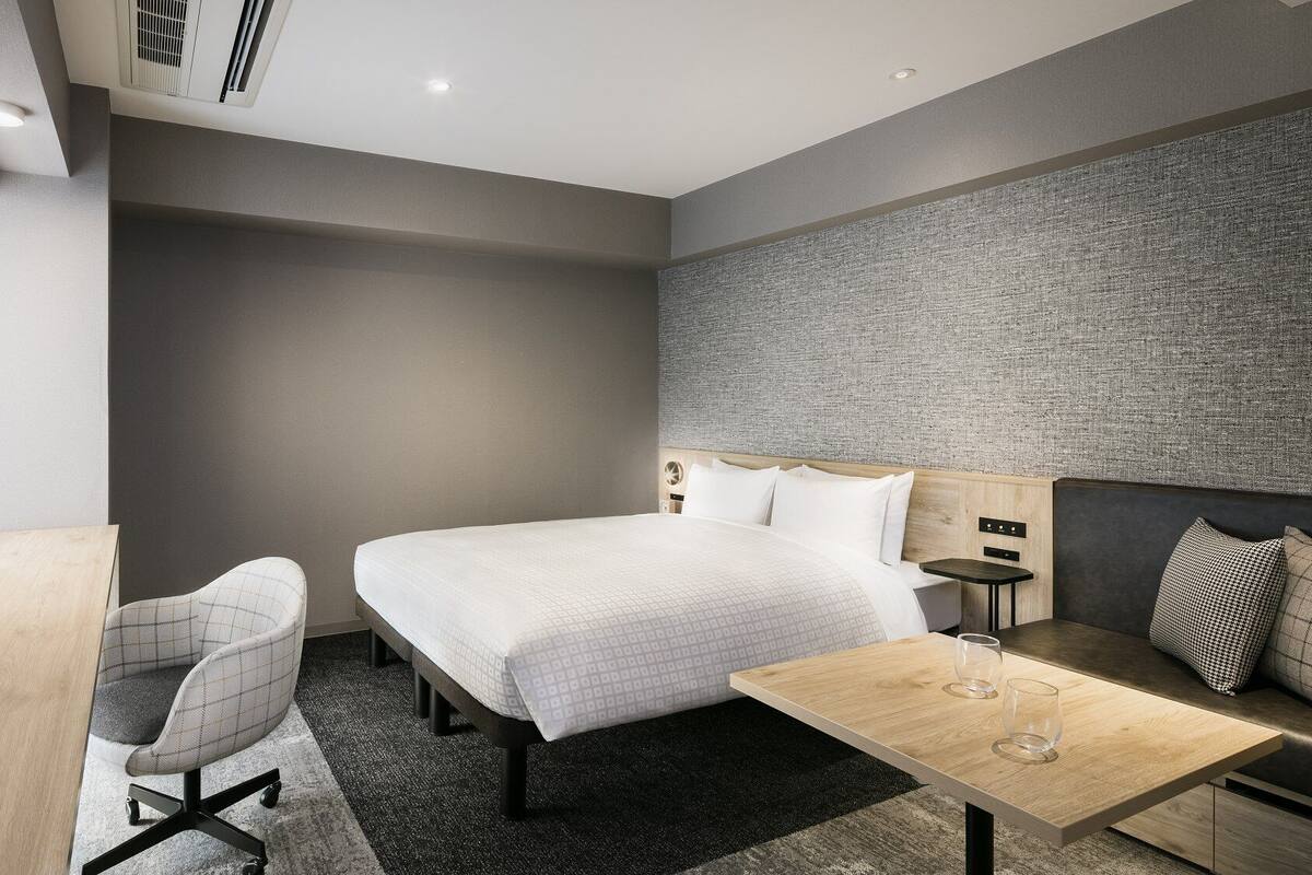 A modern hotel room with a bed, desk, chair, and a small table with a glass.