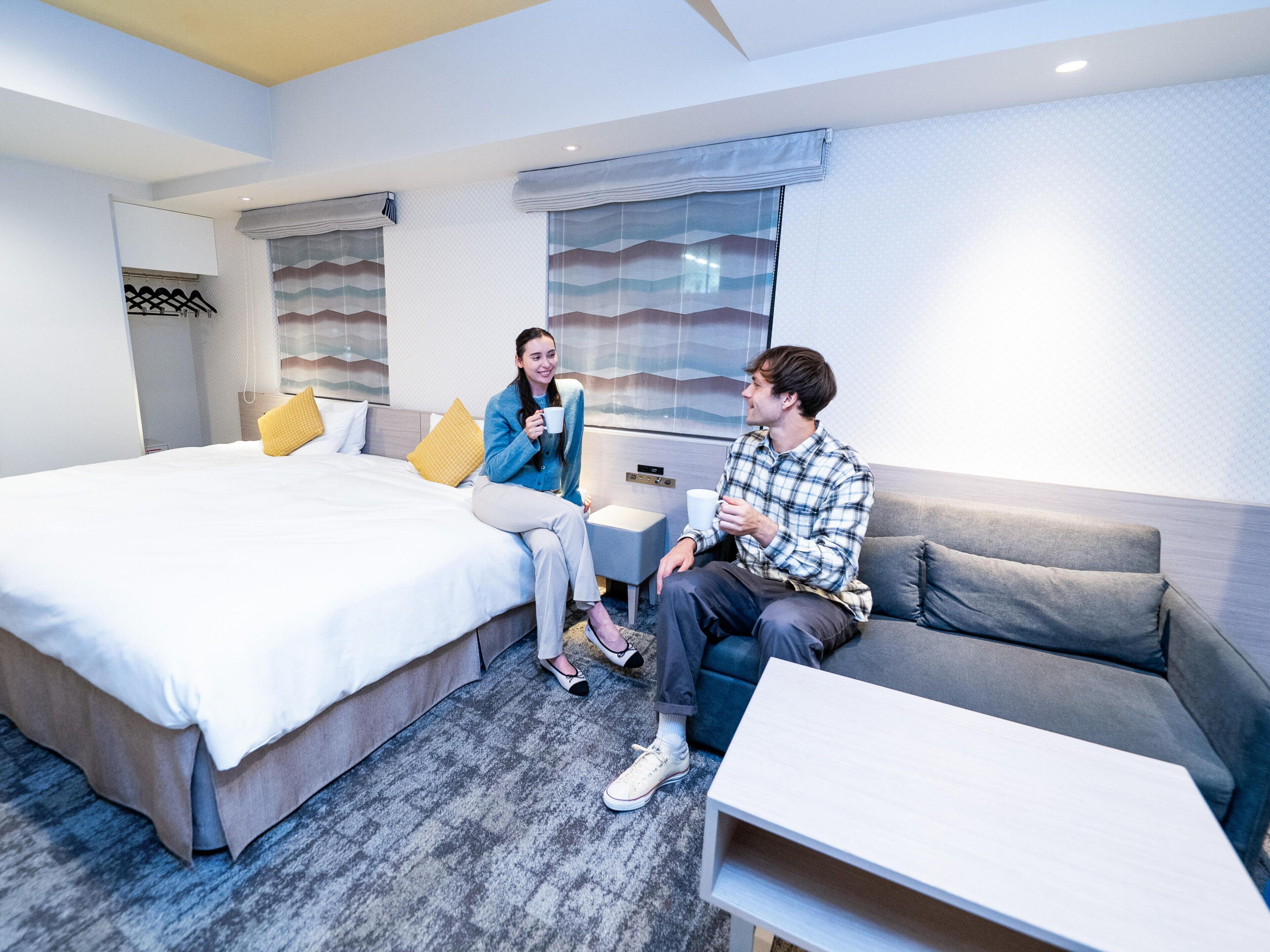 A modern hotel room with a large bed, a sofa, a small table, and a window with curtains.