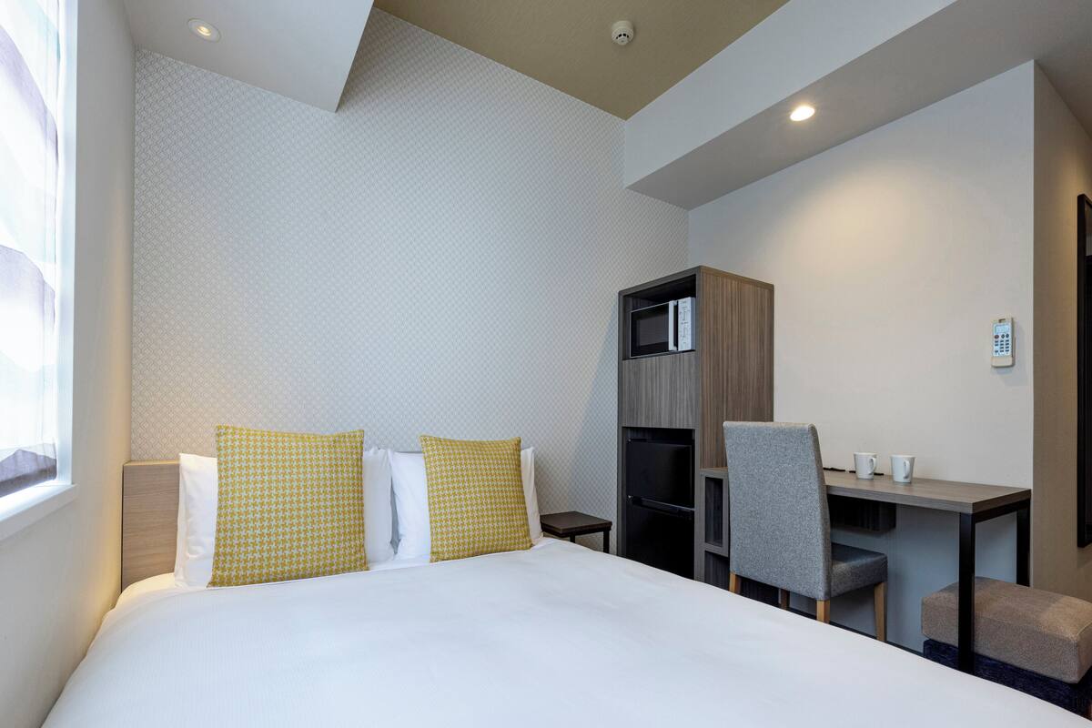 A hotel room with a bed, a desk, a microwave, and a control panel on the wall.