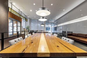 Un salón de conferencias con una larga mesa de madera, sillas blancas, un sofá de cuero marrón e iluminación colgante.