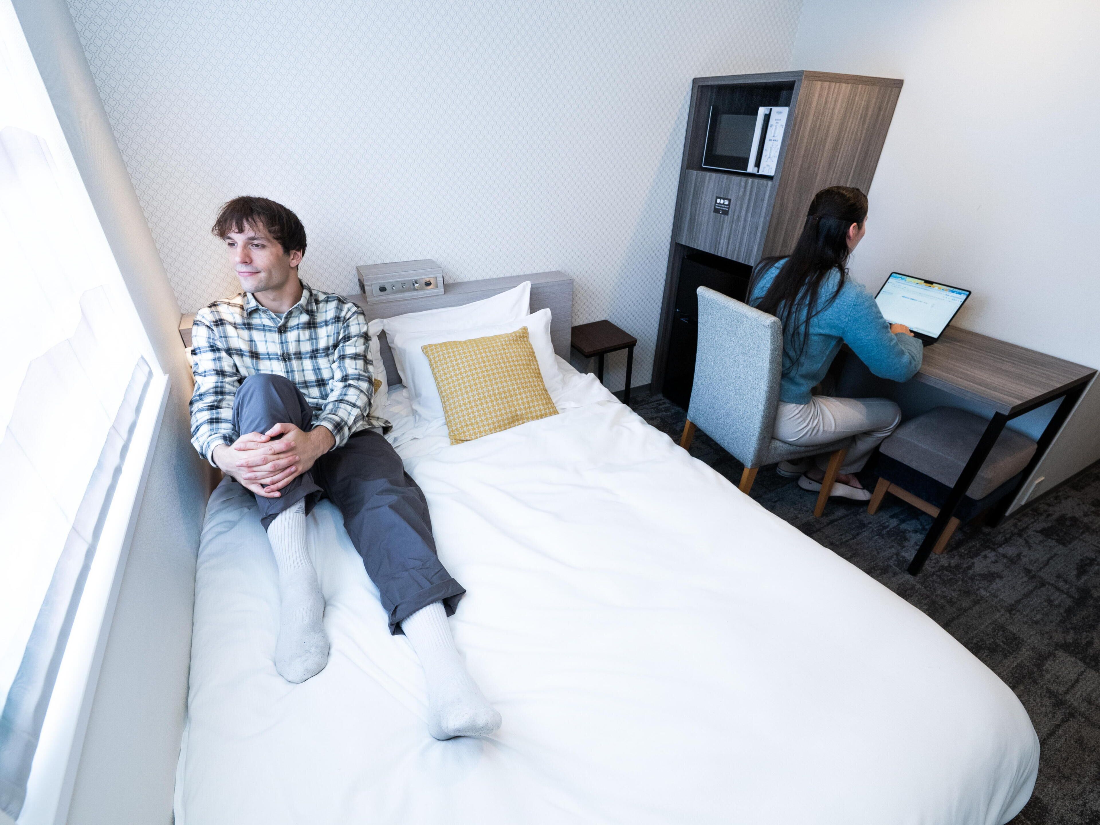 Uma cama de solteiro com colchão branco e almofada estampada, uma mesinha de cabeceira com luminária, um pequeno armário de madeira com micro-ondas e uma cadeira com laptop.