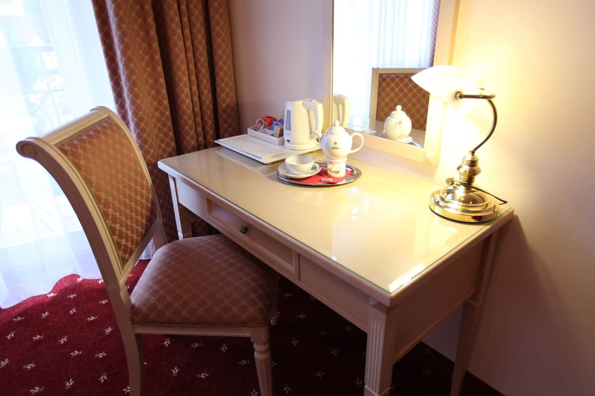 A hotel room with a white desk, a chair, a lamp, a coffee maker, and a cup.