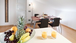 Un comedor con una mesa dispuesta para dos personas, con un frutero que incluye piña, uvas y plátanos, y dos vasos de zumo de naranja.