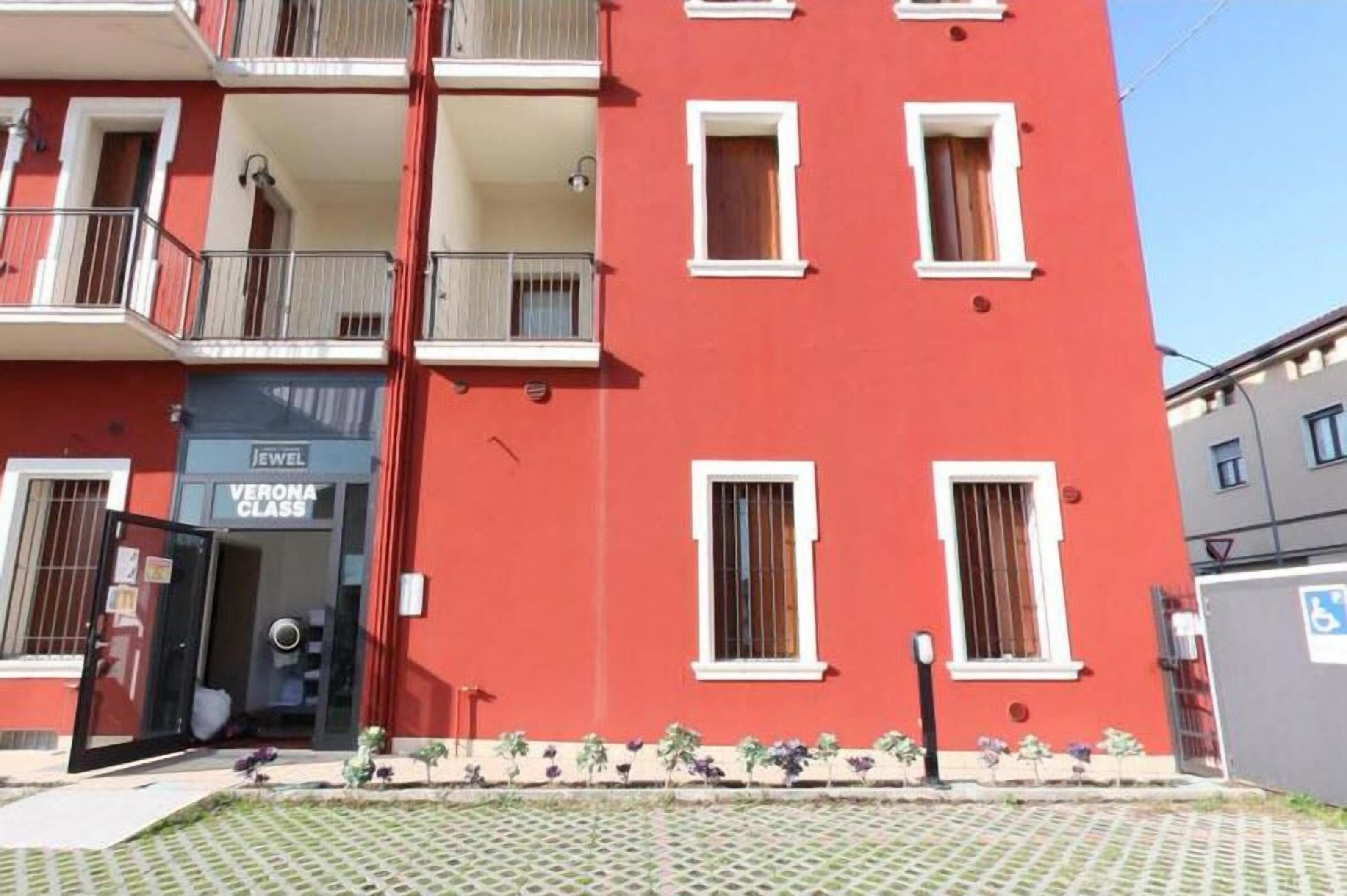 Un edificio rojo con múltiples ventanas y balcones, una entrada visible con puerta, y un pequeño jardín con plantas.