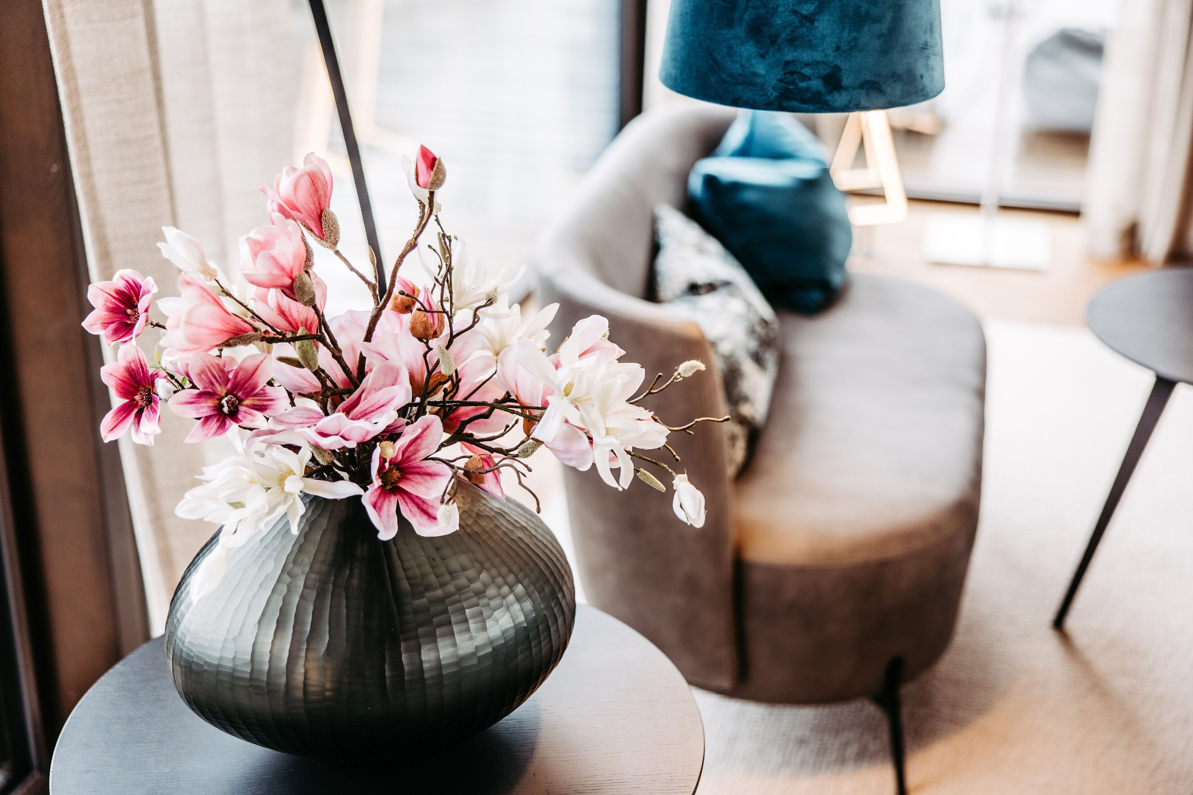 A vase with pink and white flowers on a table in a modern living room with a blue lamp and a grey sofa.