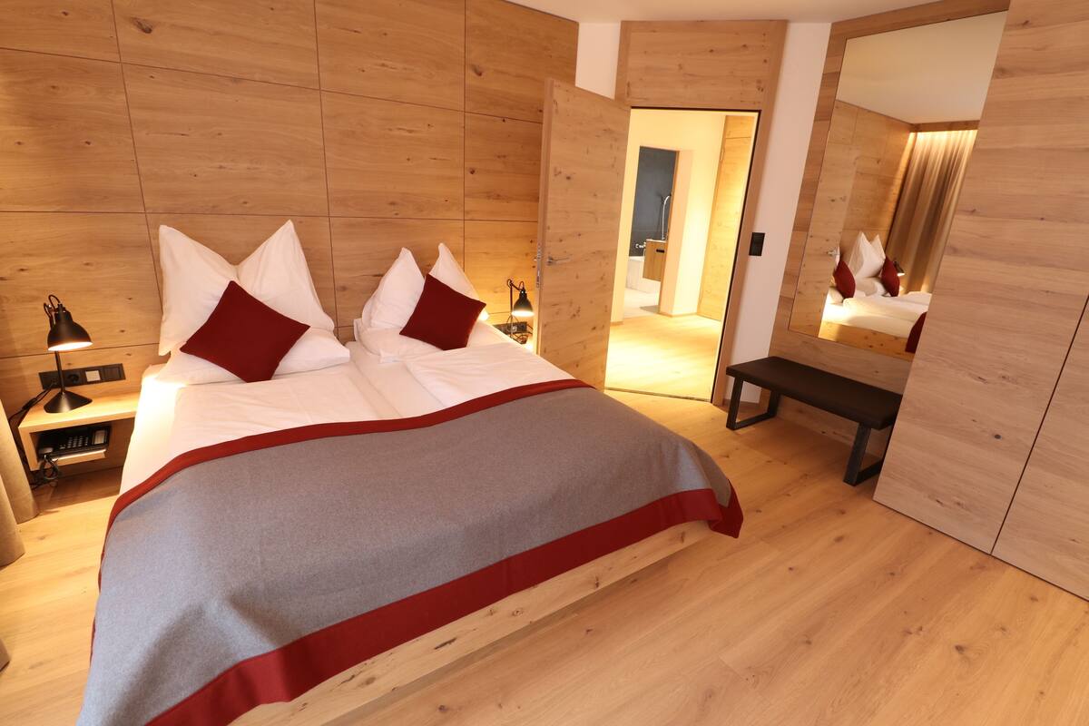 A modern bedroom with a wooden floor and walls, a large bed with white and red bedding, a bedside table with a lamp, and a bench.