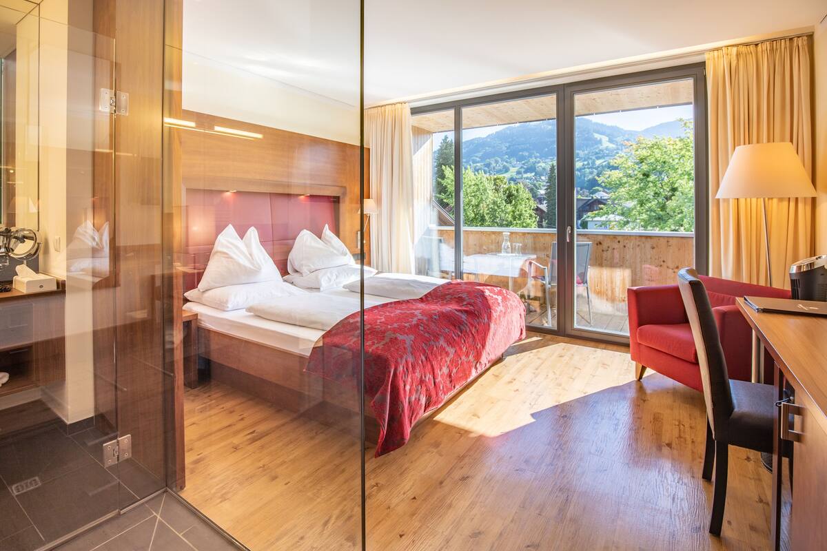 A hotel room with a bed, a desk, a chair, and a balcony with a view of the mountains.