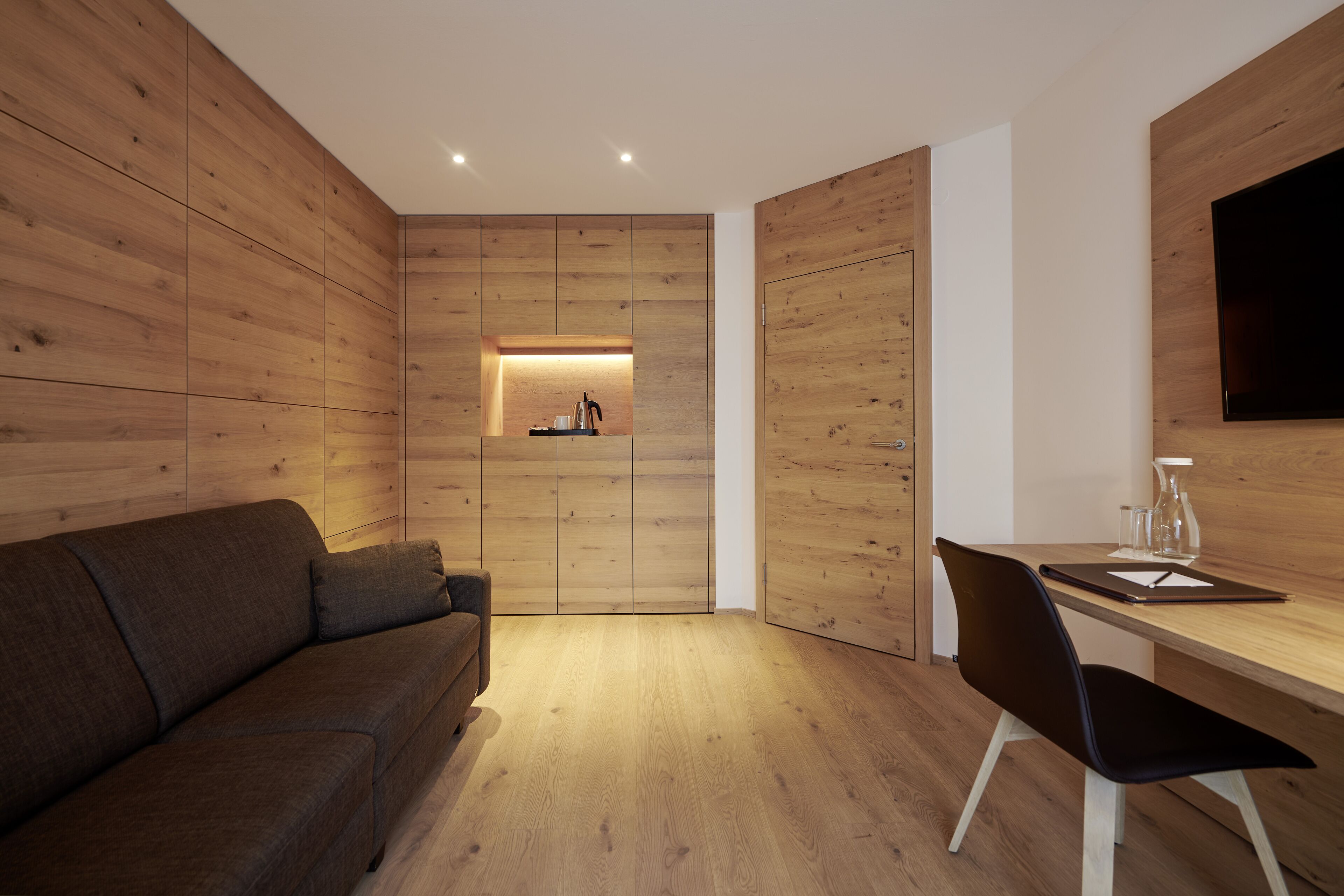 Una habitación moderna con paneles de madera, un sofá, un escritorio con una silla y un televisor de pantalla plana montado en la pared.