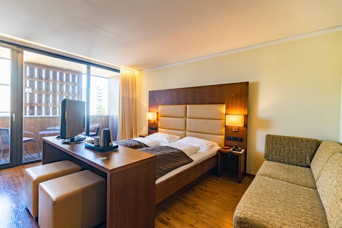 A hotel room with a bed, a desk with a computer, a chair, a sofa, and a window with curtains.