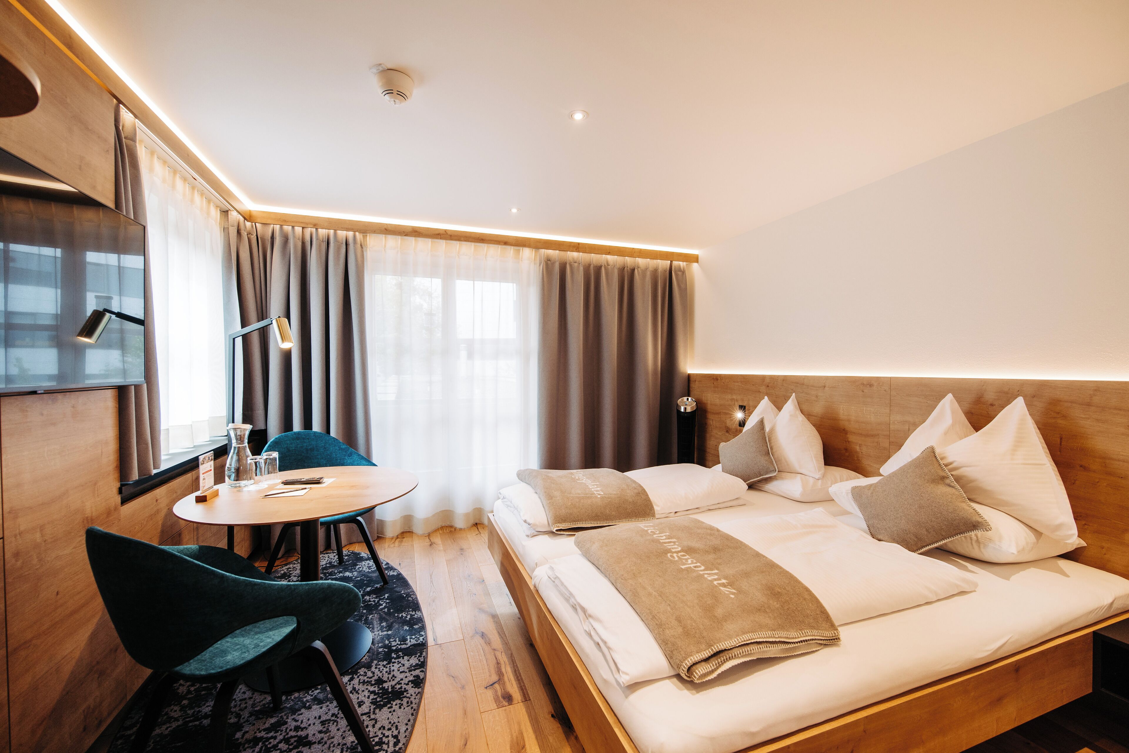 Una habitación de hotel moderna con una cama de madera, un escritorio con una silla y un ventanal con cortinas.