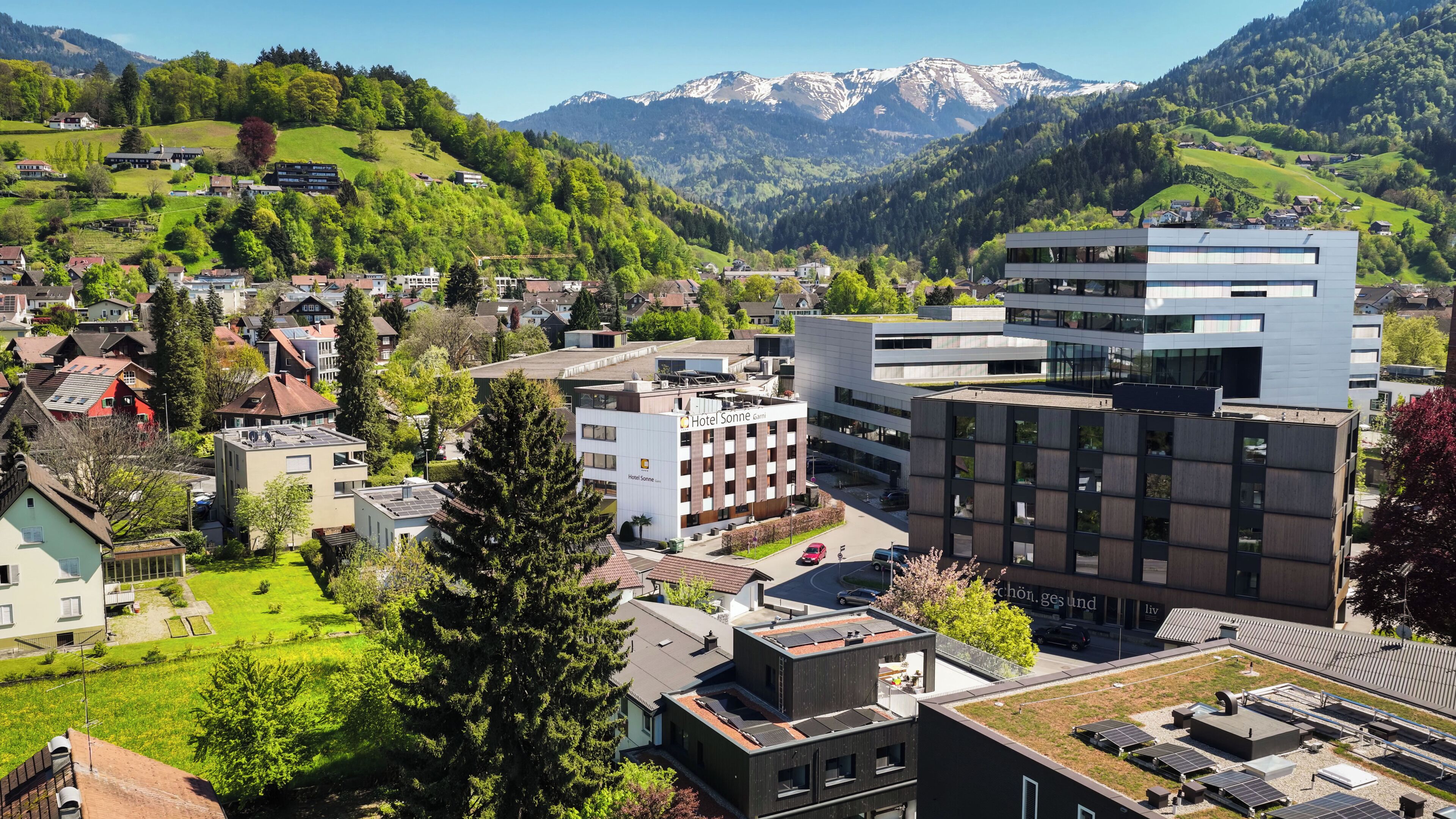 A modern hotel with a rooftop garden, surrounded by residential buildings and mountains.