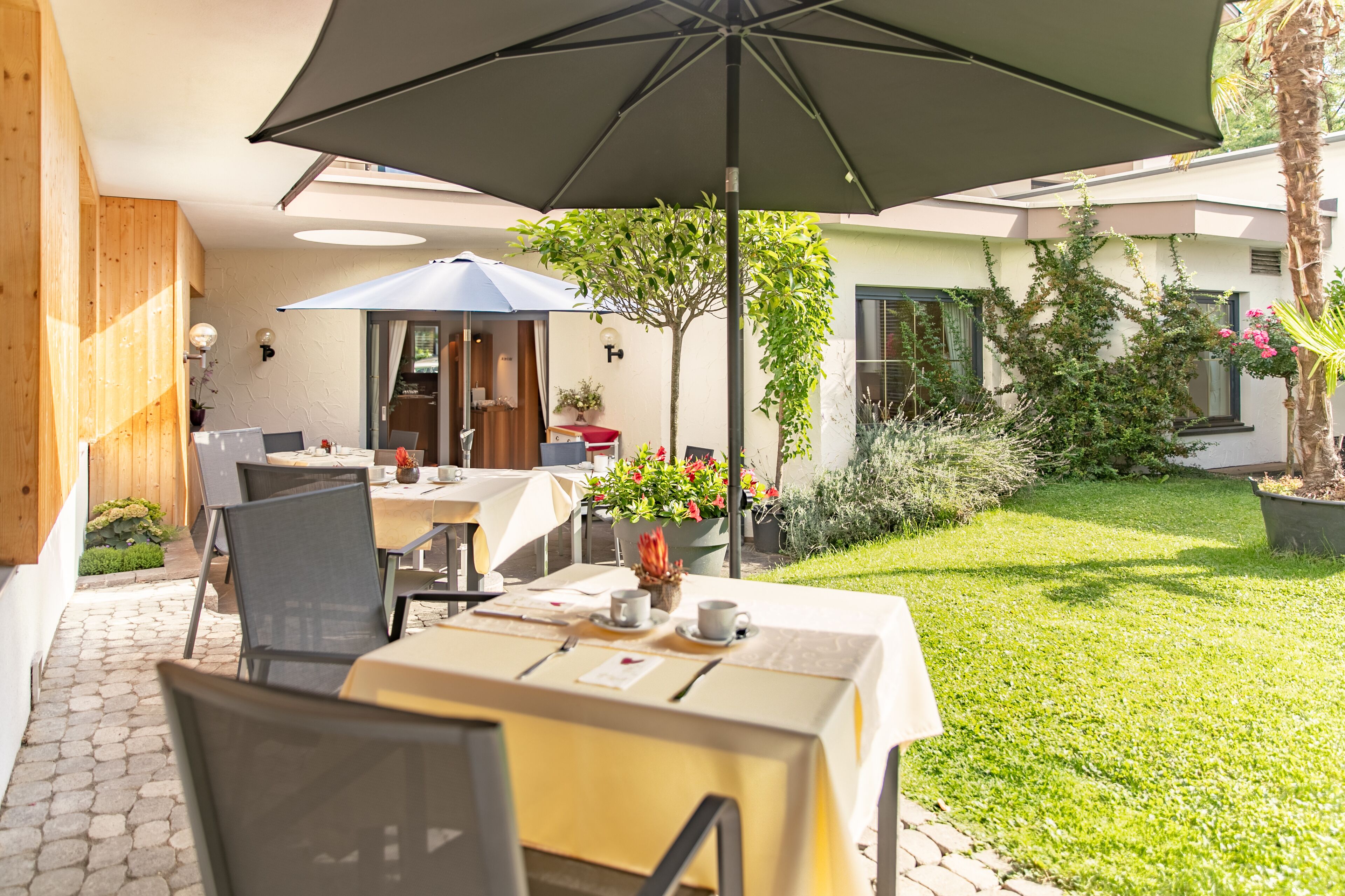 An outdoor patio with a table set for two, a large umbrella, and a garden area with a palm tree and potted plants.
