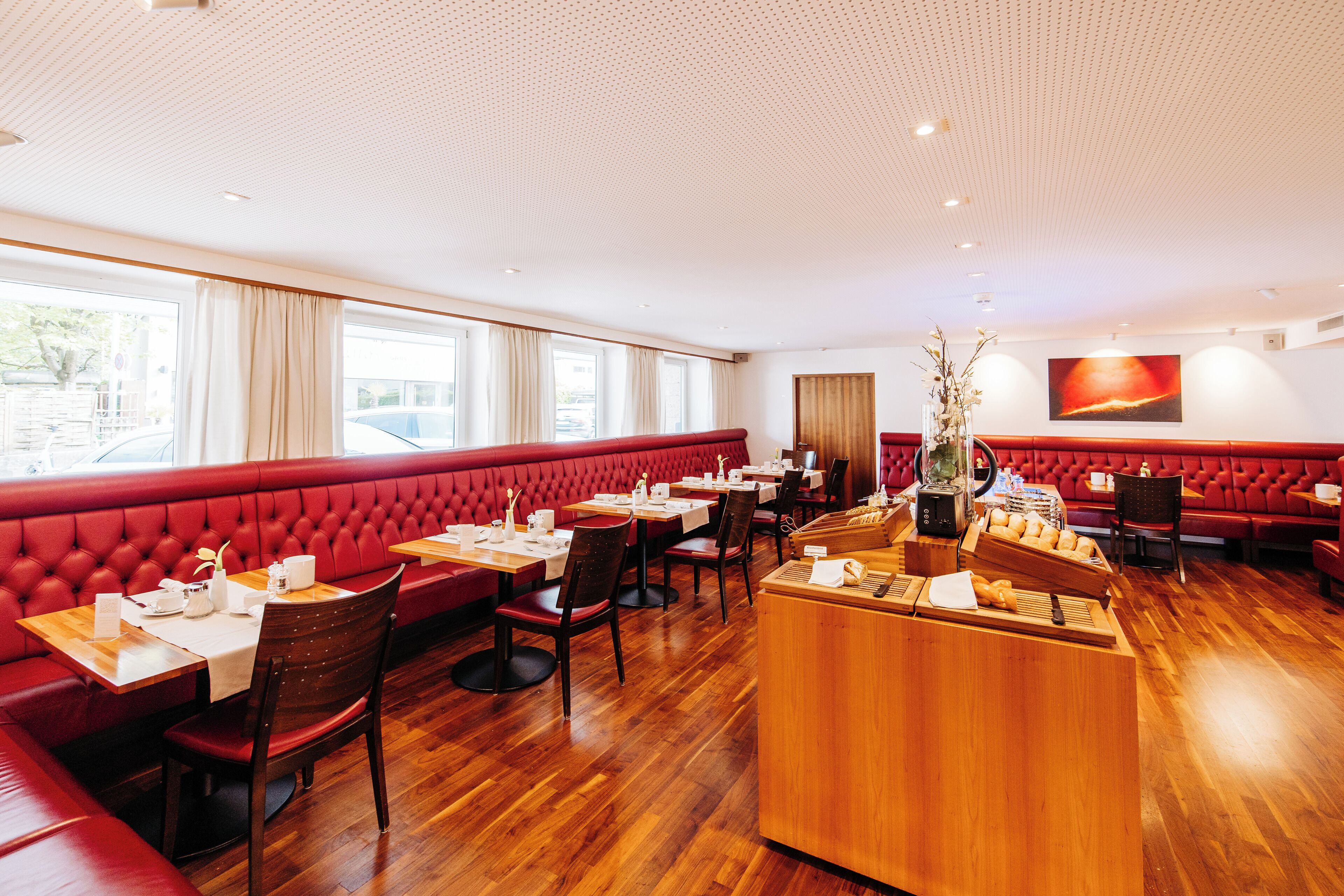 Un restaurante con asientos tapizados en rojo y mesas de madera, una barra de madera y un amplio ventanal con cortinas.