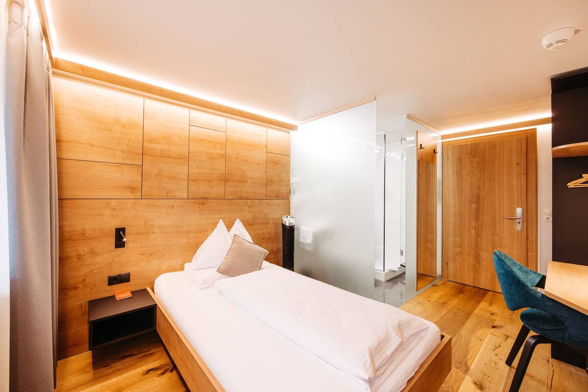 A modern hotel room with a wooden headboard, a bed with white linens, a desk with a chair, and a glass shower enclosure.