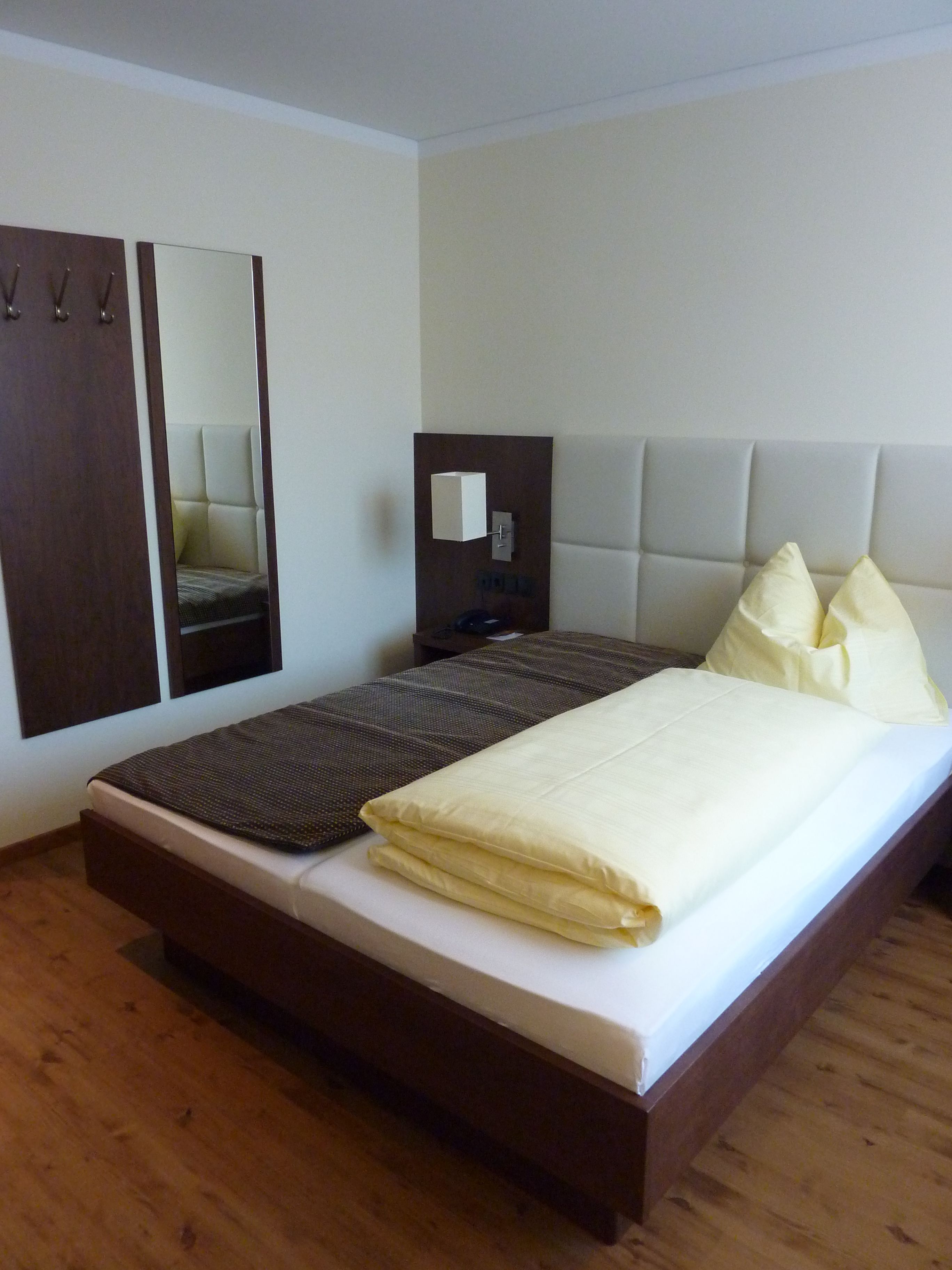 Ein ordentlich bezogenes Bett mit weißer Matratze und braunem Kopfteil. An der Wand links vom Bett befindet sich ein großer Spiegel.