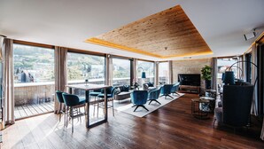 Amplia sala de estar con techo de madera, ventanas grandes que ofrecen vistas al campo, comedor con sillas azules y televisor sobre un mueble.