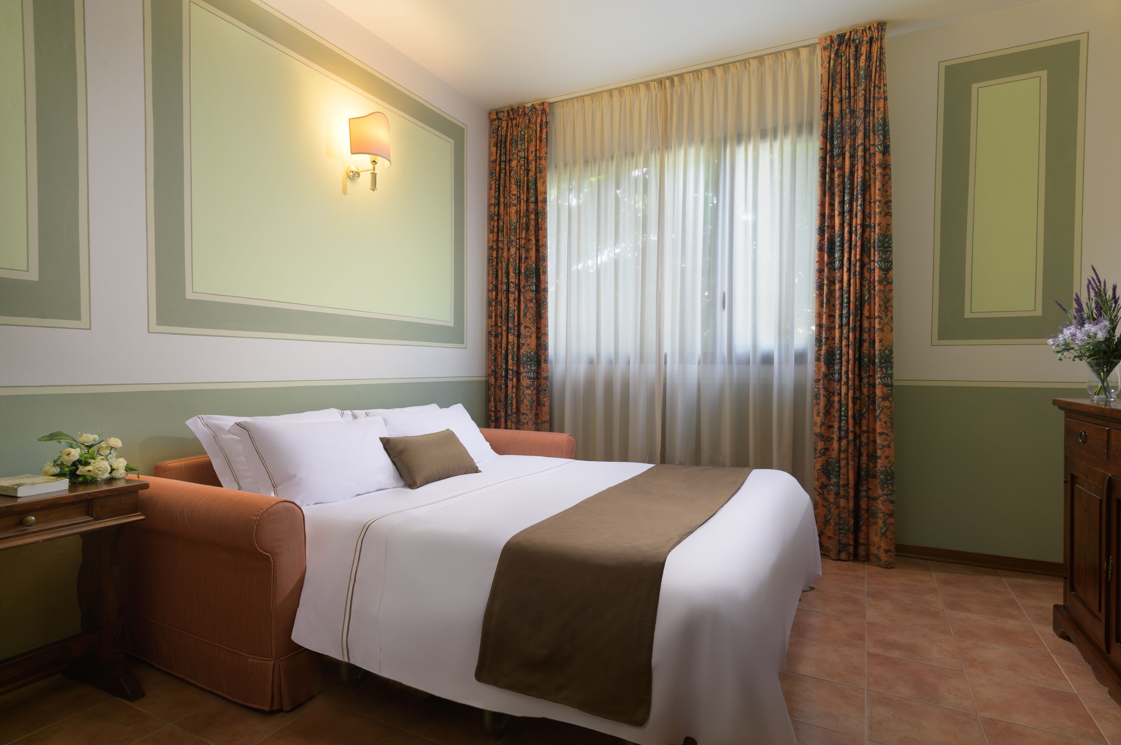 Una cama bien hecha con una colcha blanca y marrón, una manta marrón y una sola almohada. A la izquierda de la cama hay una mesita de noche con una lámpara y un jarrón con flores. La habitación tiene un ventanal con cortinas traslúcidas y una cortina estampada a la derecha.