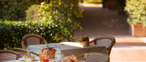 Un espacio de comedor al aire libre con sillas de mimbre, una mesa servida con pan, fruta y una bebida.