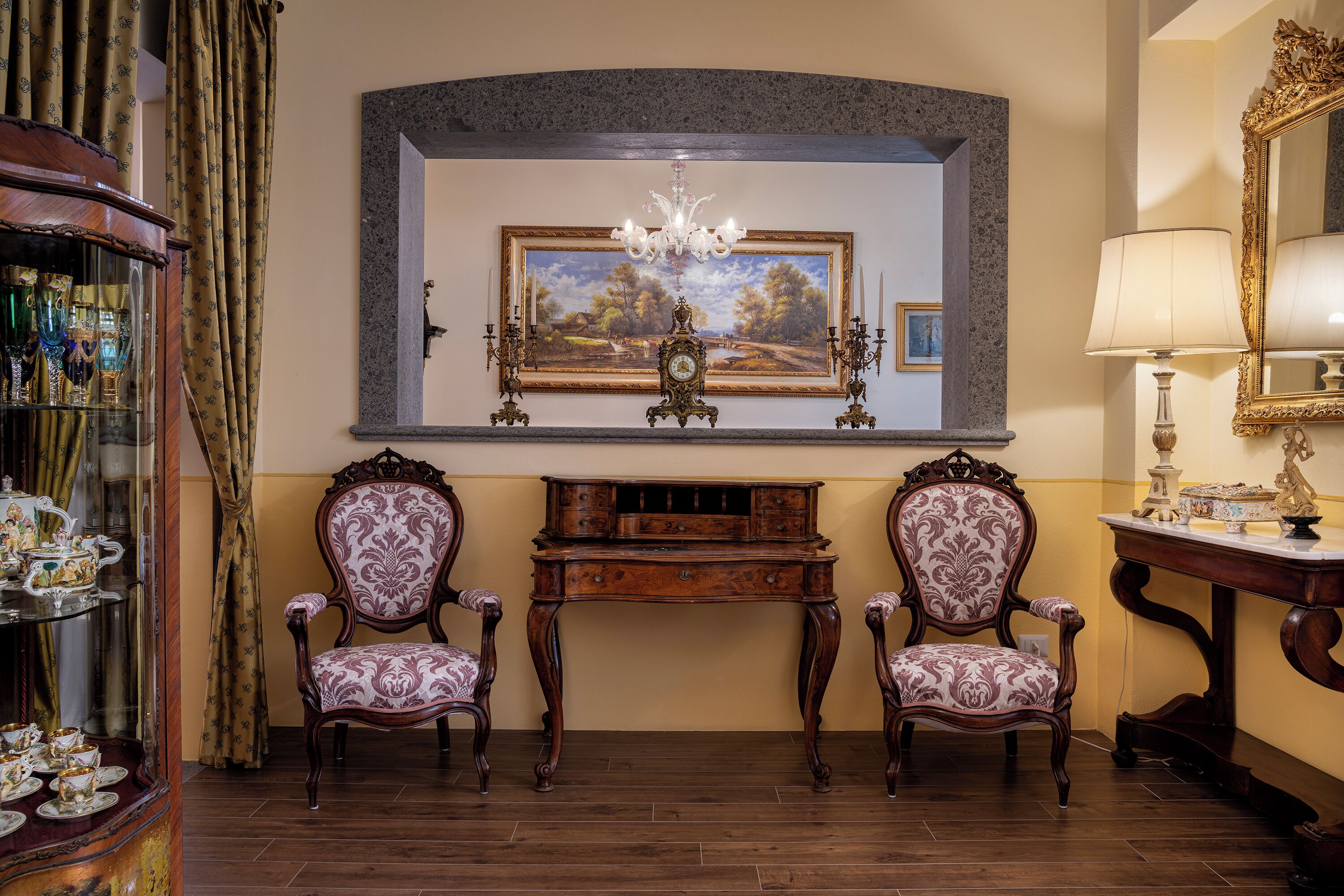 Une pièce comprenant deux fauteuils richement décorés, un buffet en bois aux portes vitrées, un tableau accroché au-dessus d’une console et un miroir au mur.