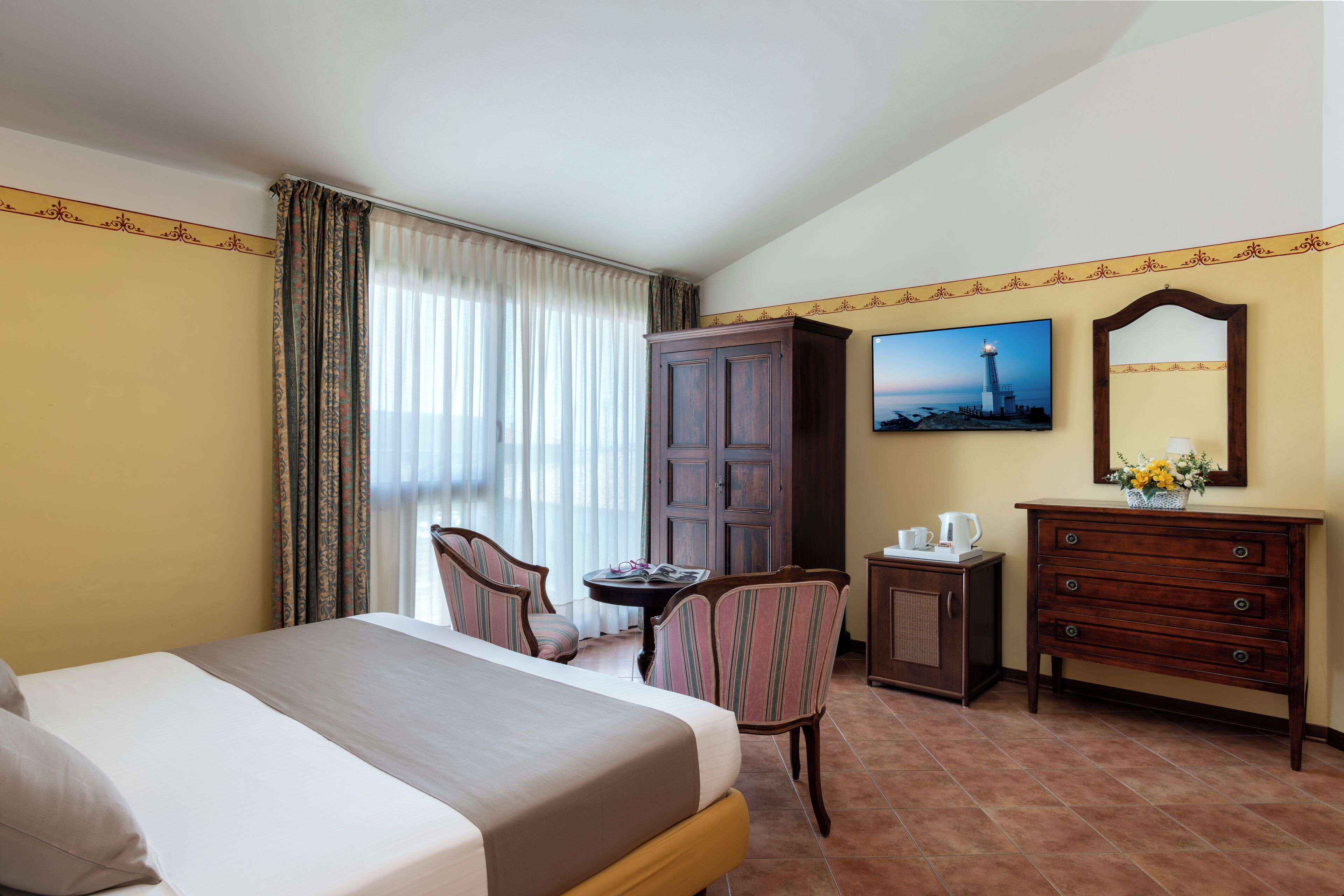 Quarto de hotel com cama, escrivaninha, cadeira, cômoda, televisão e janela com cortinas.