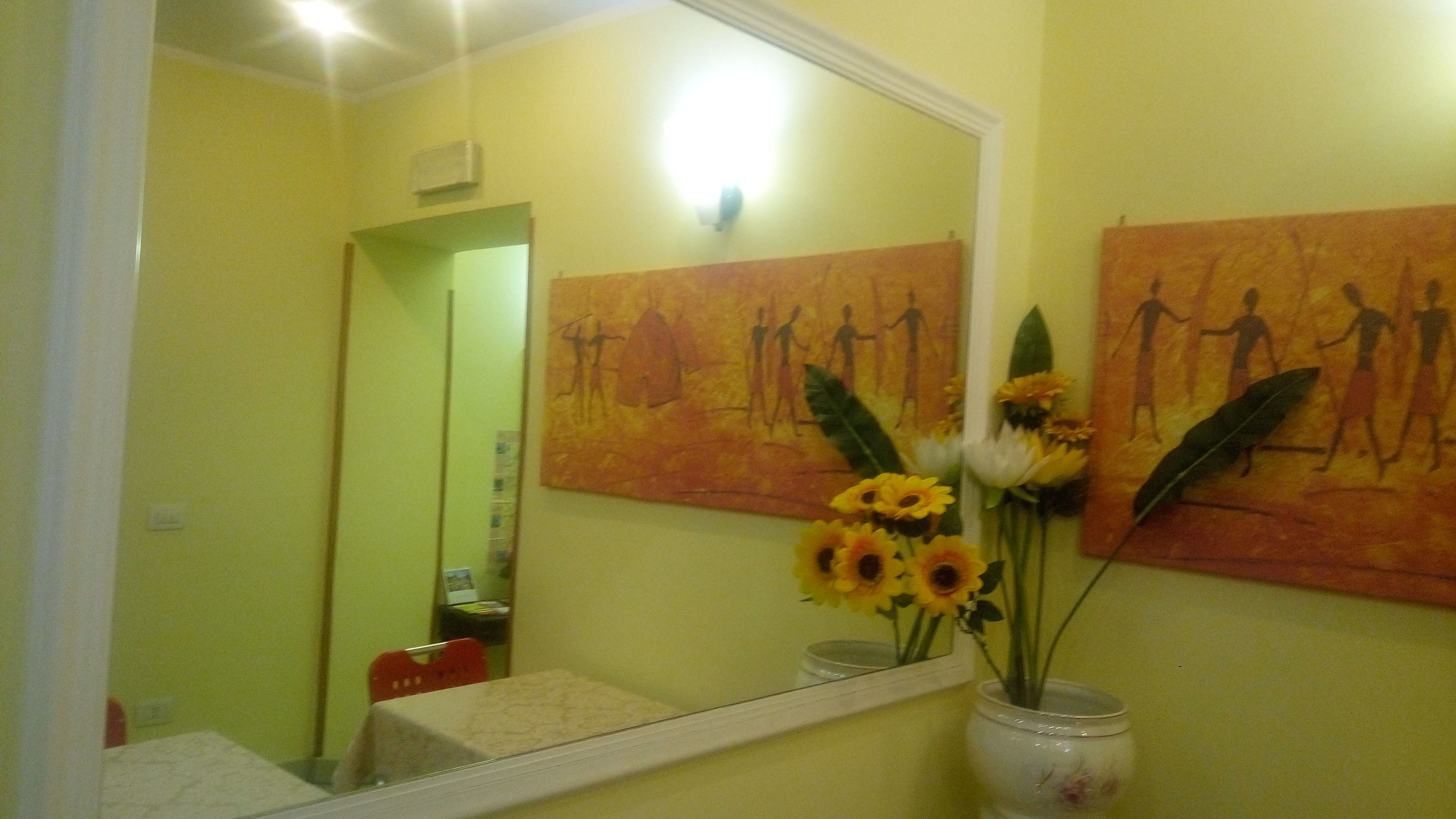 A bathroom with yellow walls, a large mirror, and artwork depicting human figures. There is a vase with sunflowers on the counter.