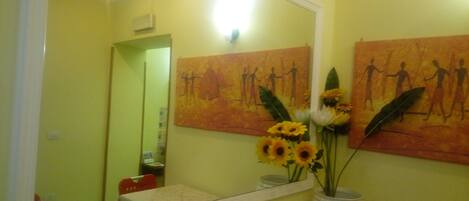 A bathroom with yellow walls, a large mirror, and artwork depicting human figures. There is a vase with sunflowers on the counter.