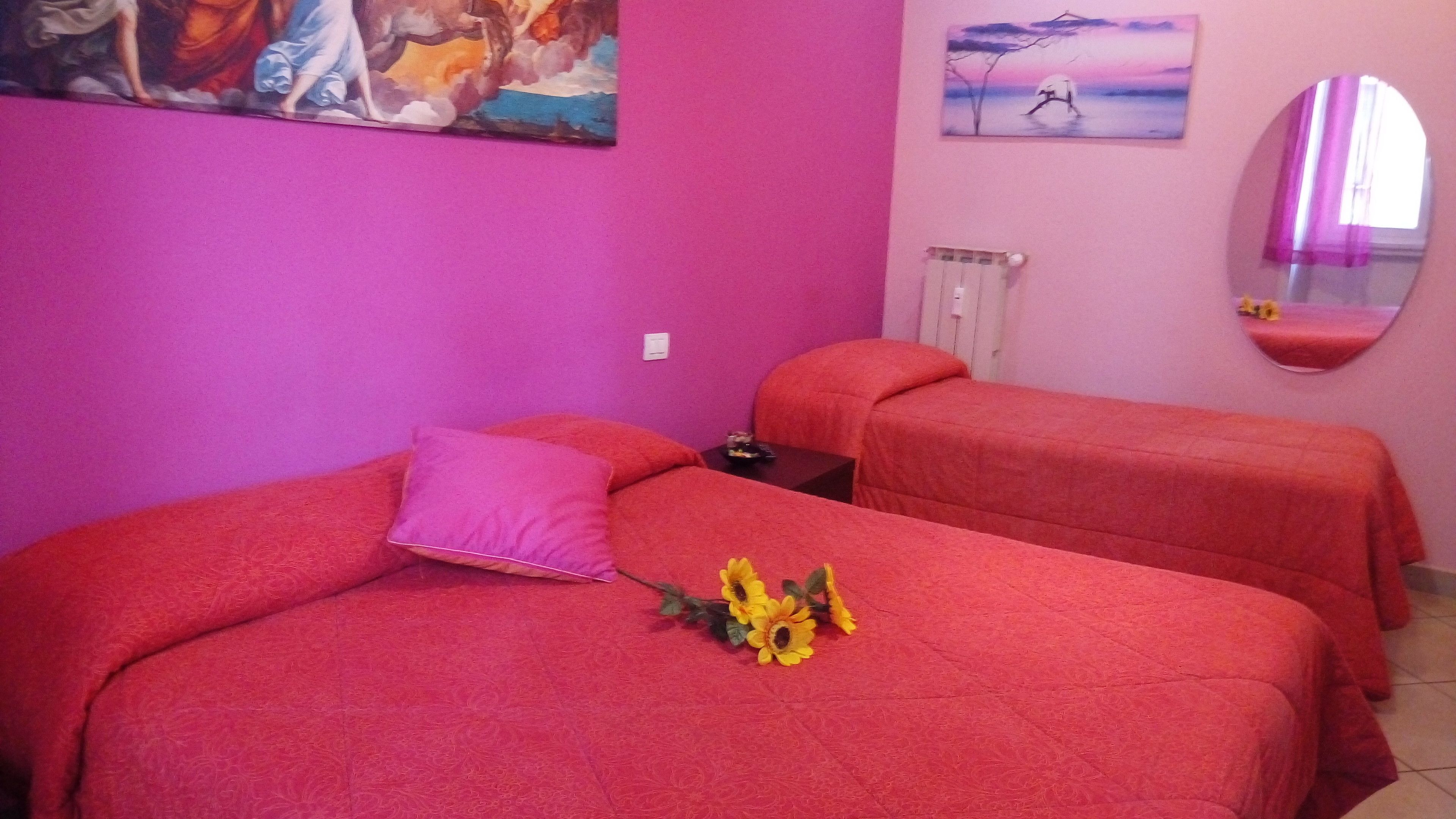 Quarto com duas camas, cada uma com colcha vermelha e um único travesseiro. Há uma pequena mesa com uma luminária e alguns objetos sobre ela. Um quadro está na parede, e um espelho na parede oposta.