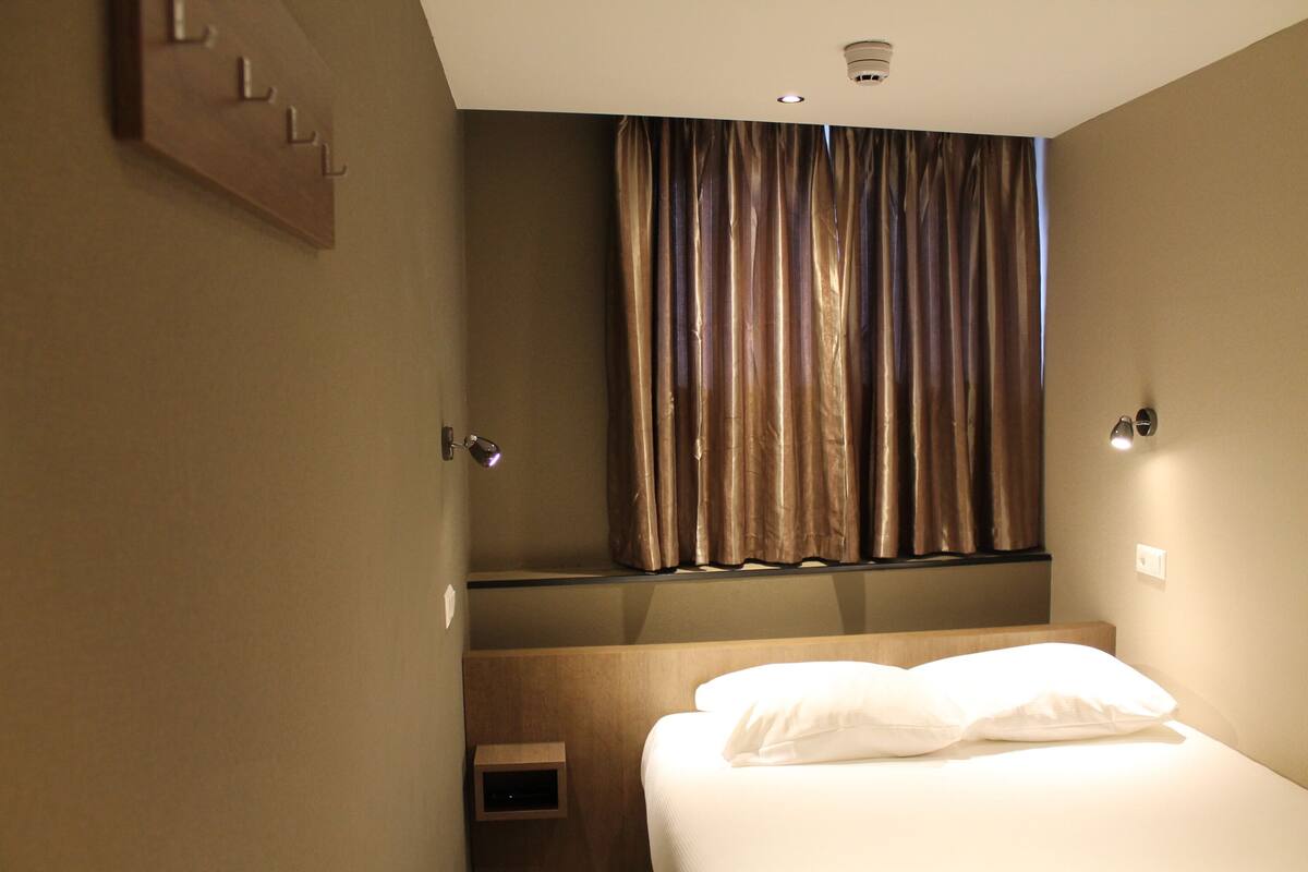 A hotel room with a bed, a headboard, a wall-mounted shelf, and a window with curtains.