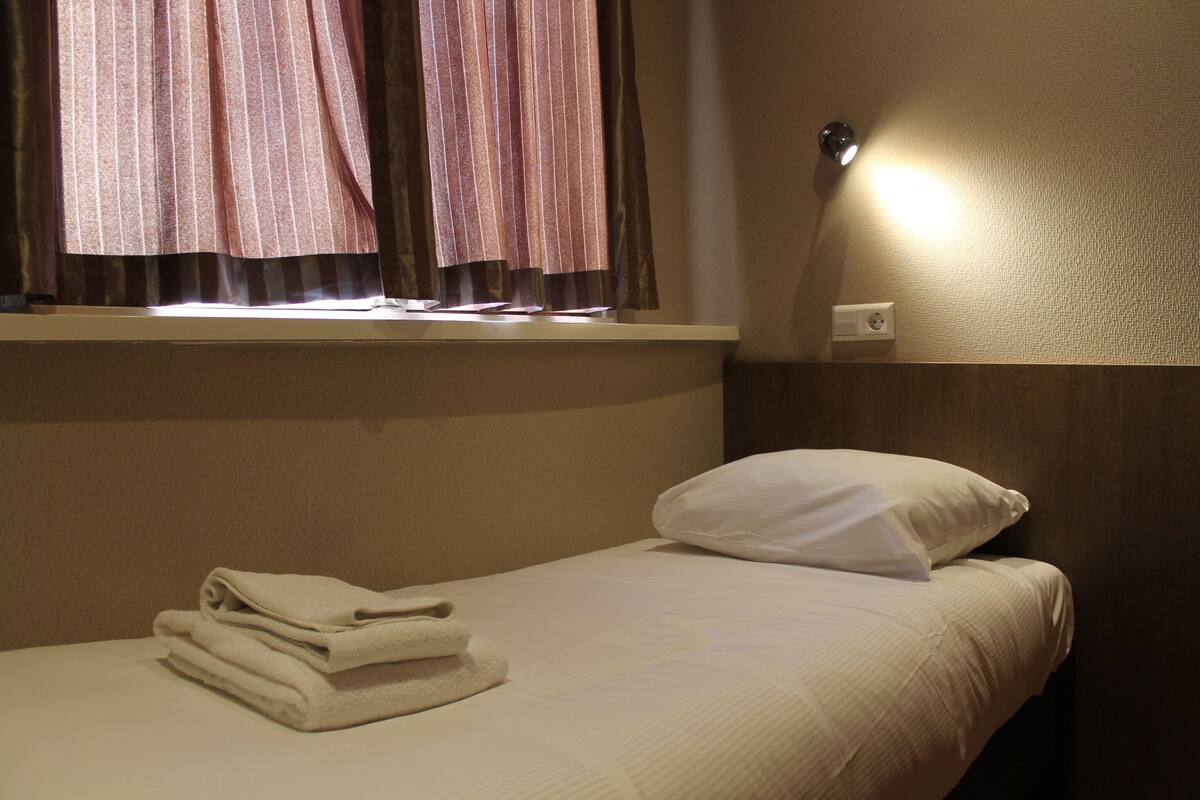 A hotel room with a bed, a window with curtains, a wall-mounted light, and a folded towel on the bed.