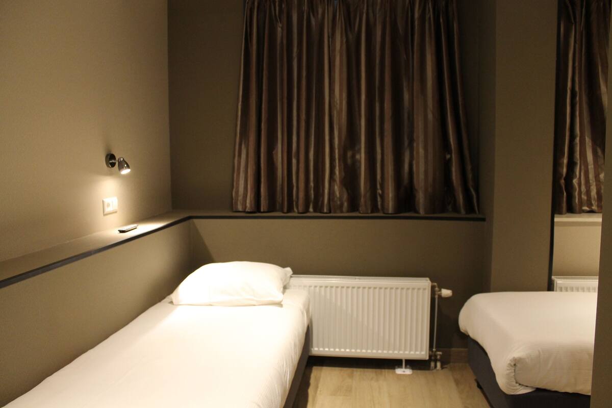 A hotel room with two single beds, a window with curtains, a wall-mounted light, and a radiator.