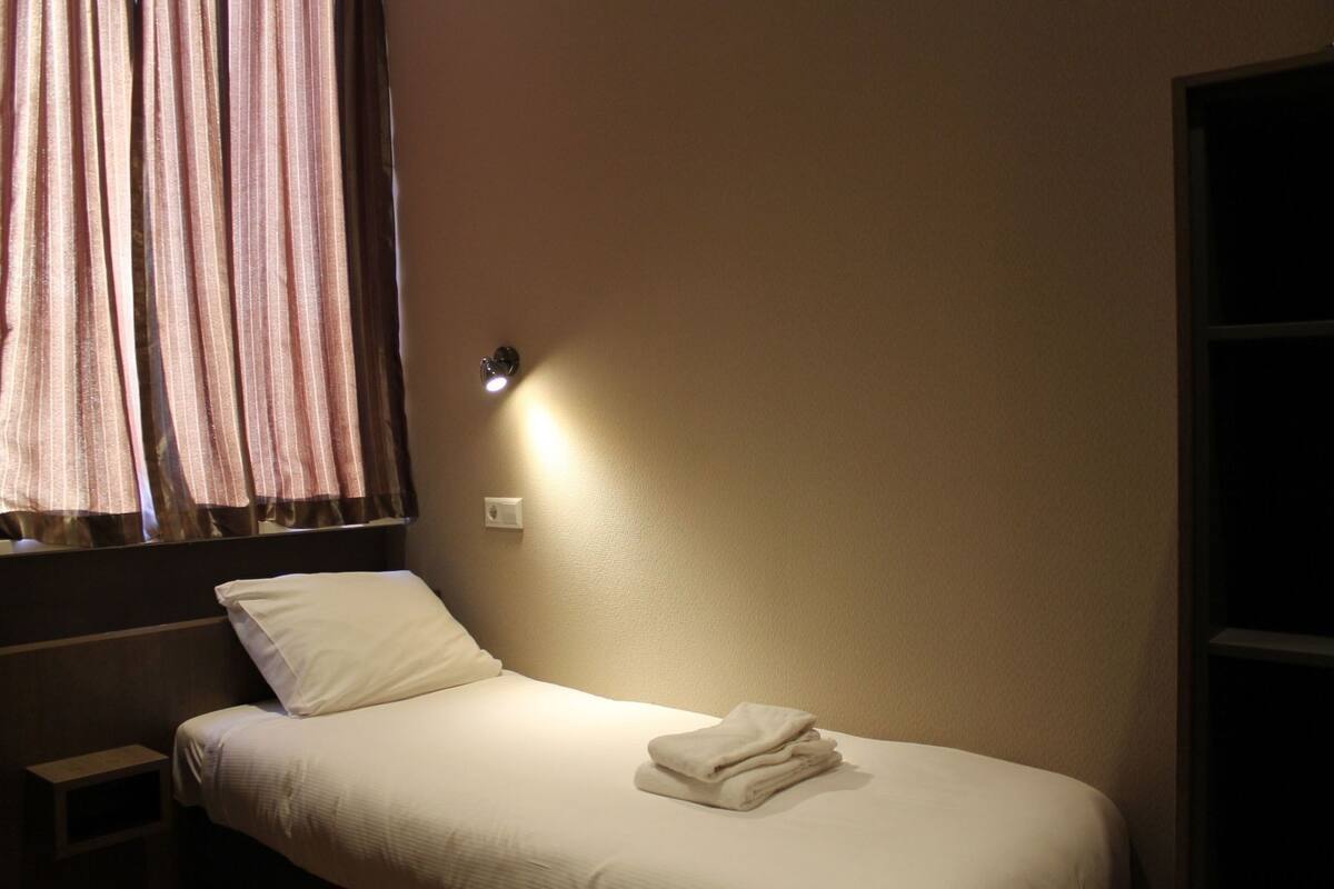 A single bed with a white mattress and pillow, a small bedside table, a wall-mounted light, and a window with curtains.