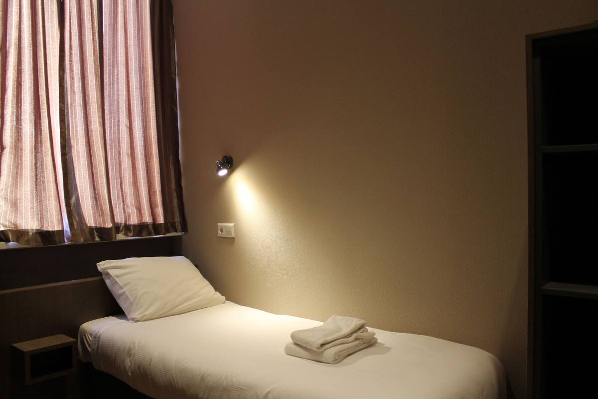 A single bed with a folded white towel, a bedside table, and a wall-mounted light.